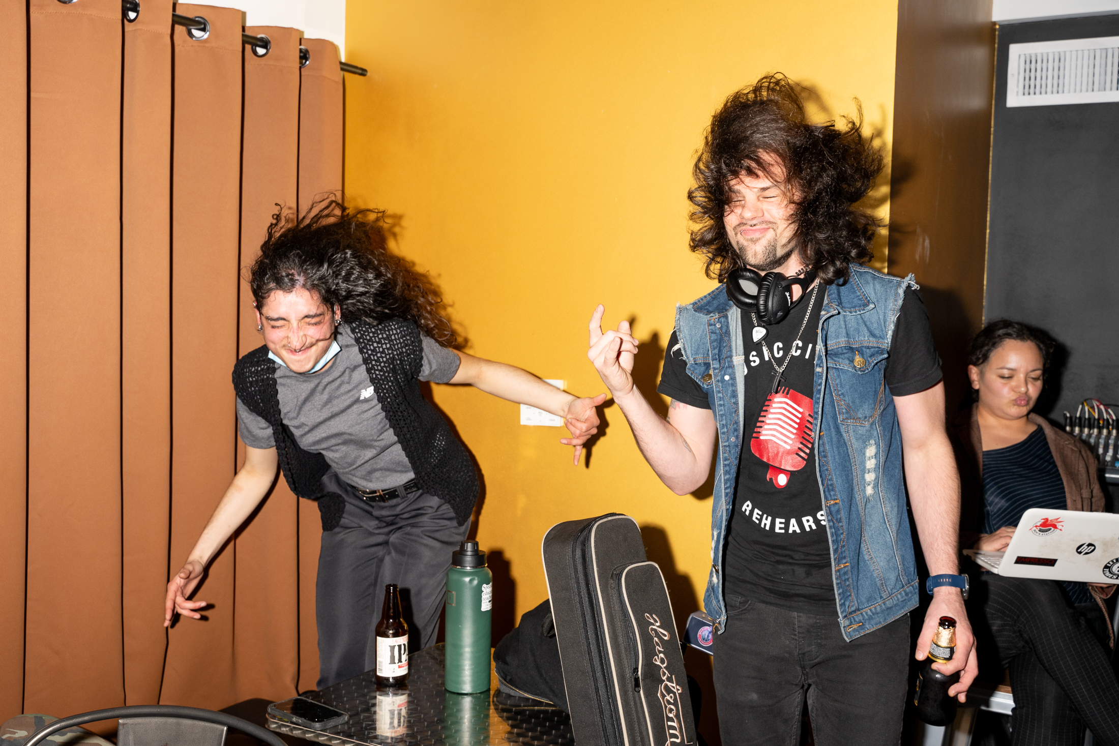 Two people are energetically dancing, hair flying, in front of a curtain. One holds a beer, the other a guitar case. A person sits in the background with a laptop.