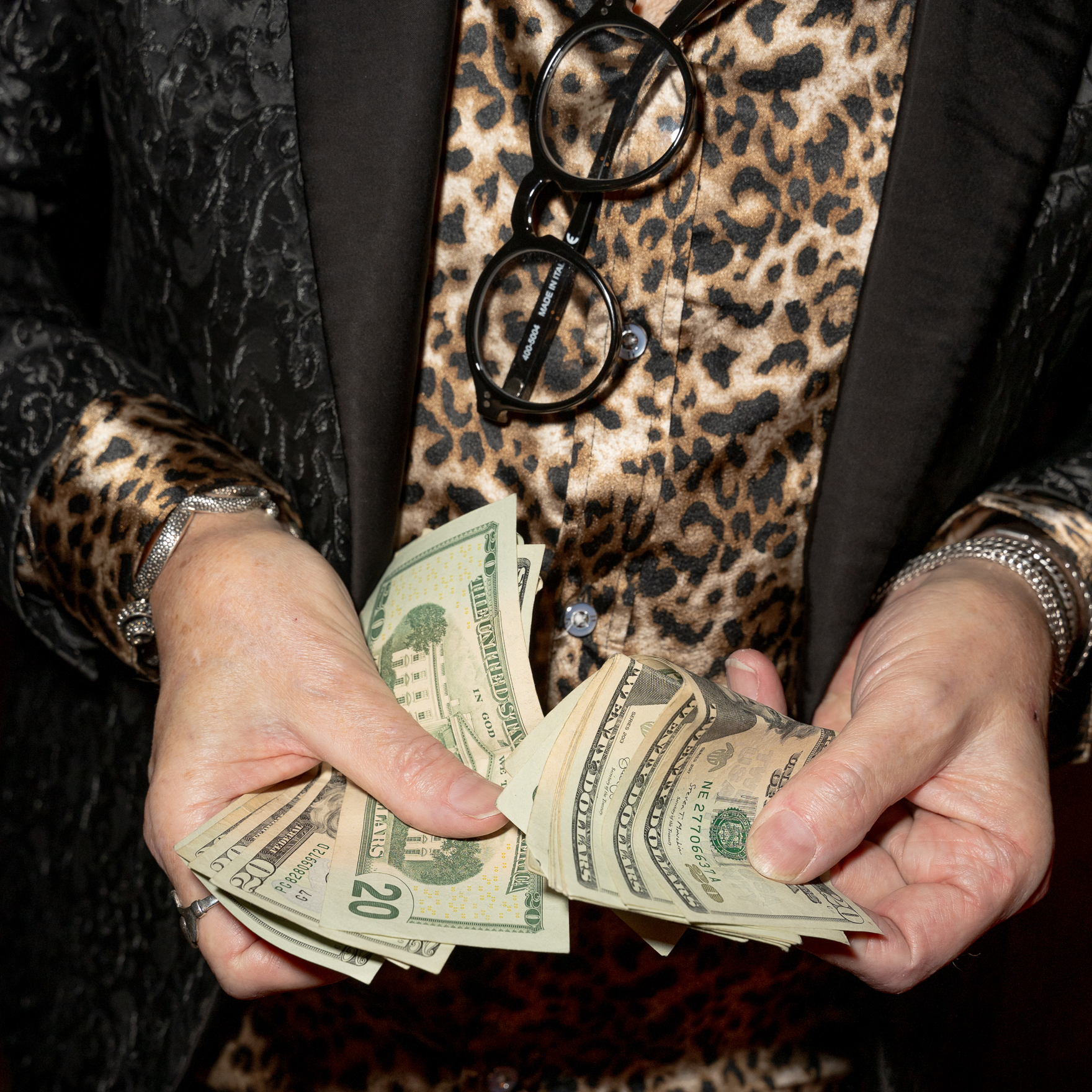 A person wearing a leopard print shirt and black patterned jacket holds a stack of U.S. dollar bills. They have round glasses and a silver bracelet.