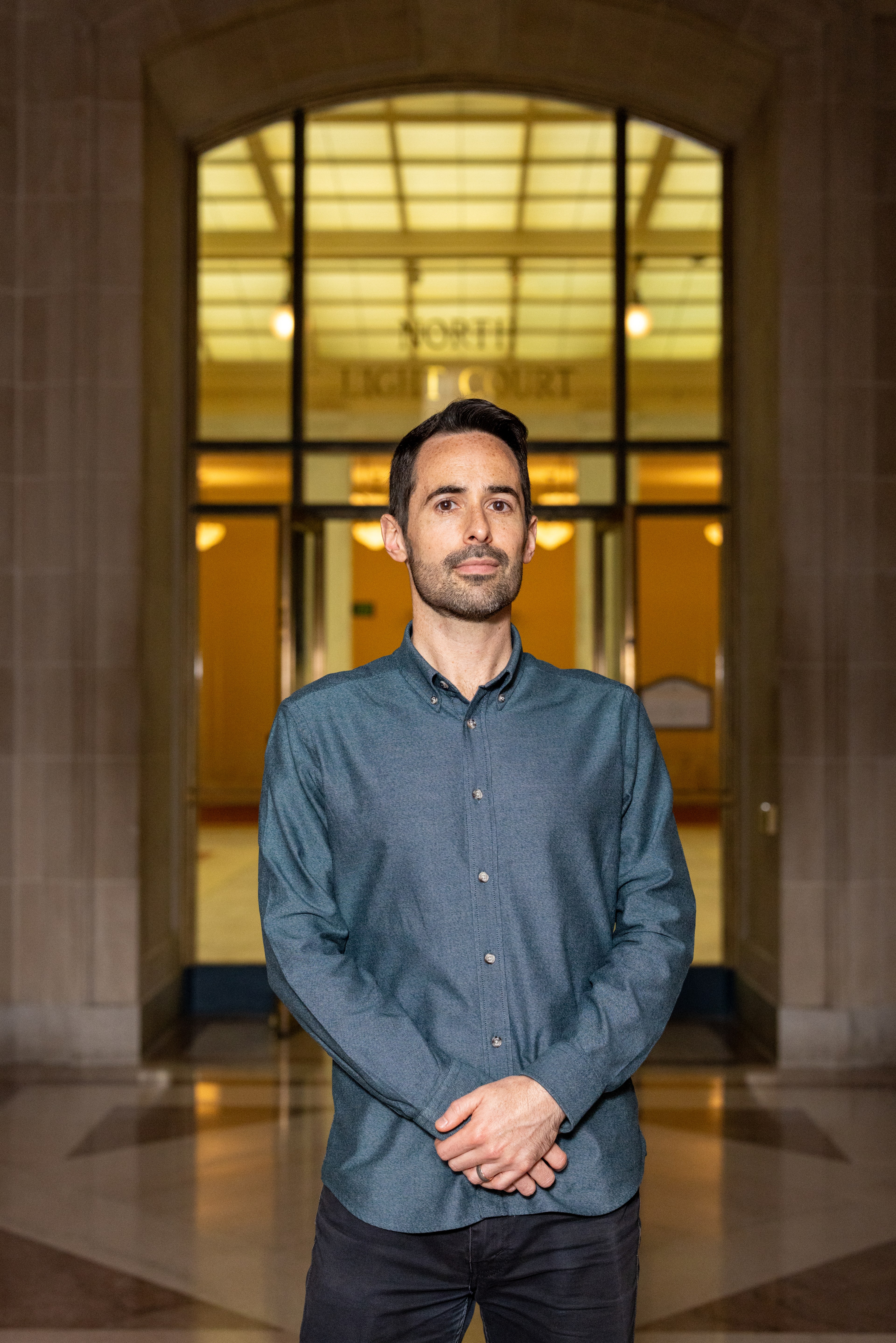 A man in a teal button-up shirt stands indoors before a large glass door labeled &quot;North Light Court.&quot; The area is softly lit with warm ceiling lights.