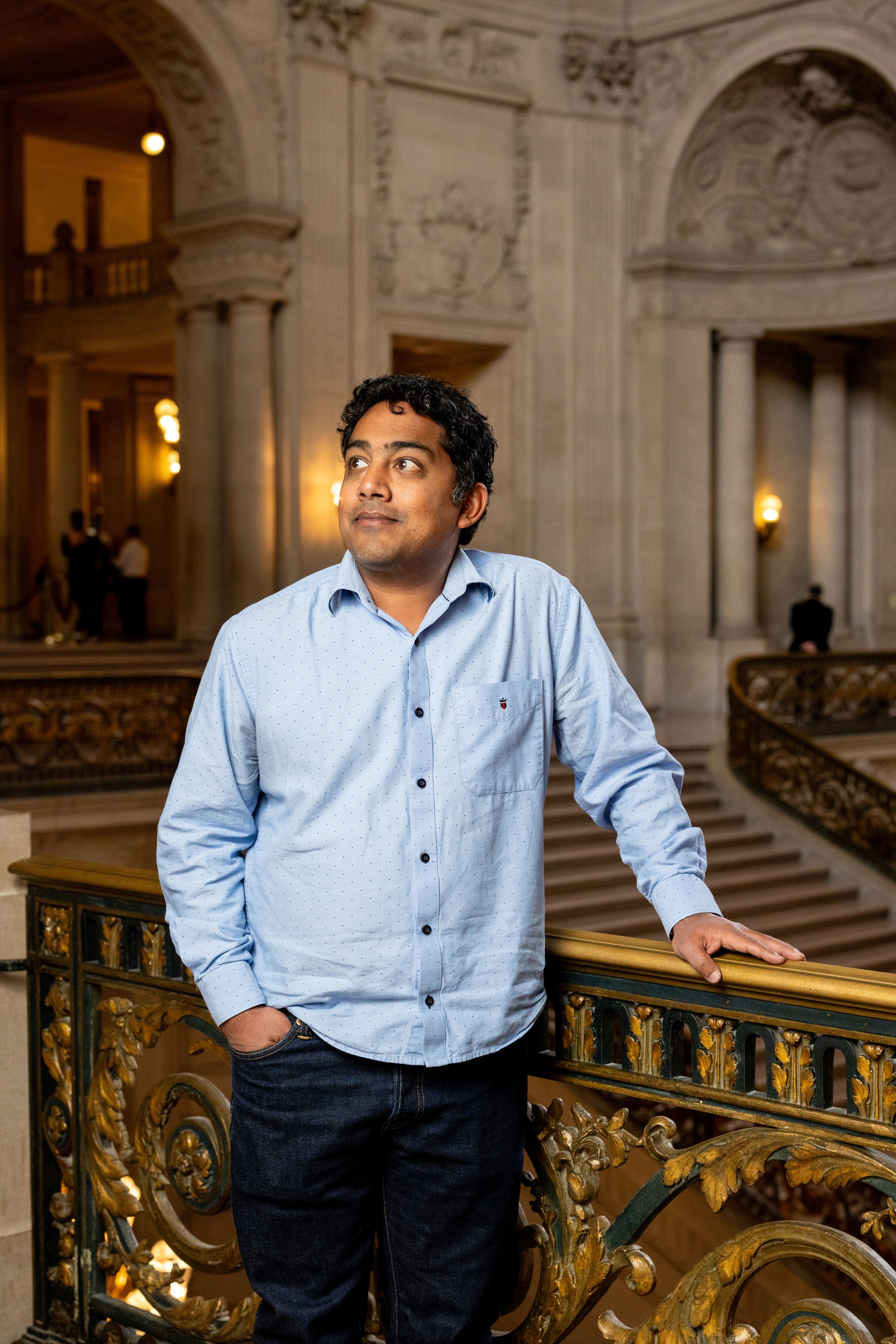 A man in a light blue shirt leans on a decorative railing inside an ornate building with high ceilings and elegant arches.