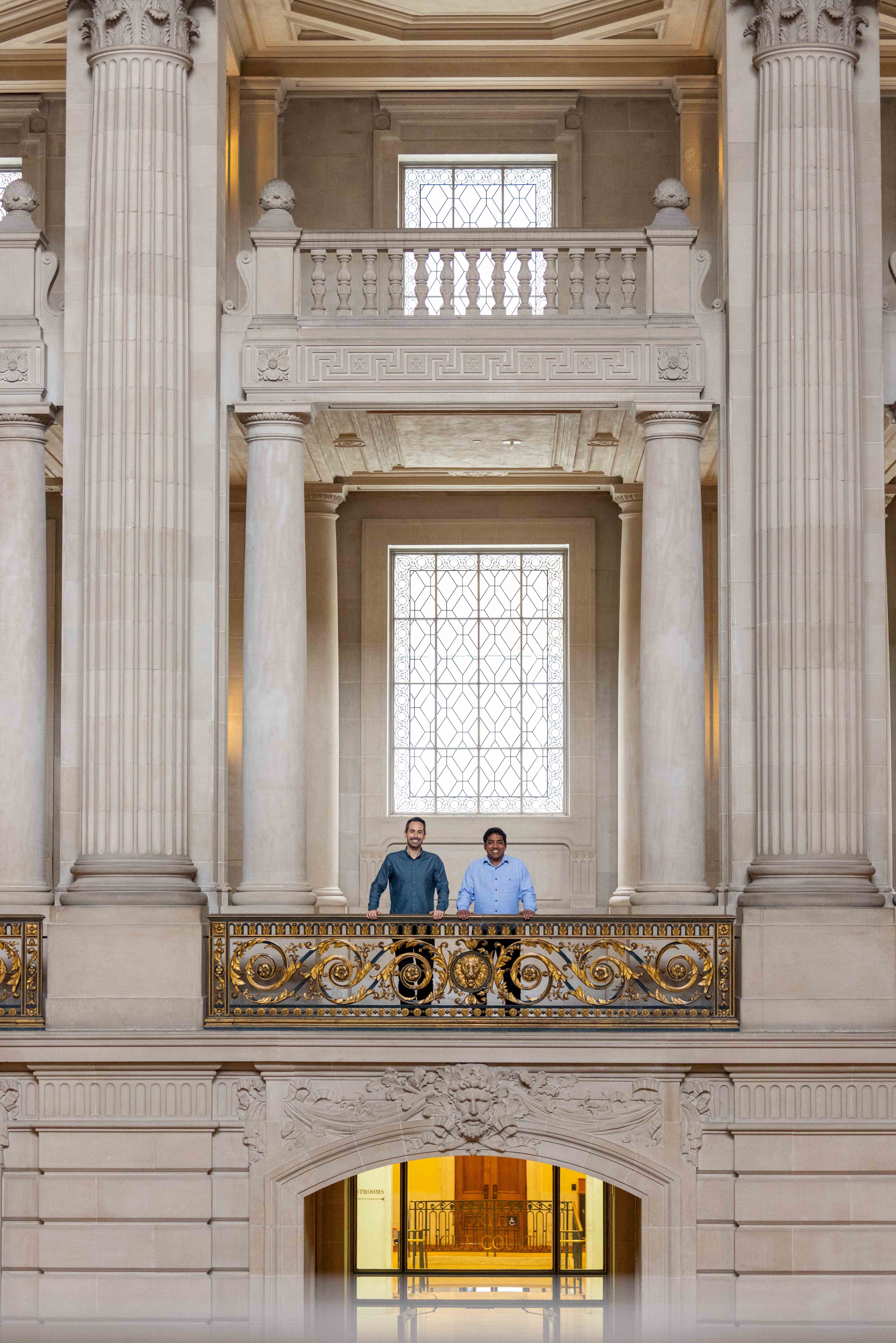 Two people stand on an ornate balcony in a grand building with large columns and decorative windows.