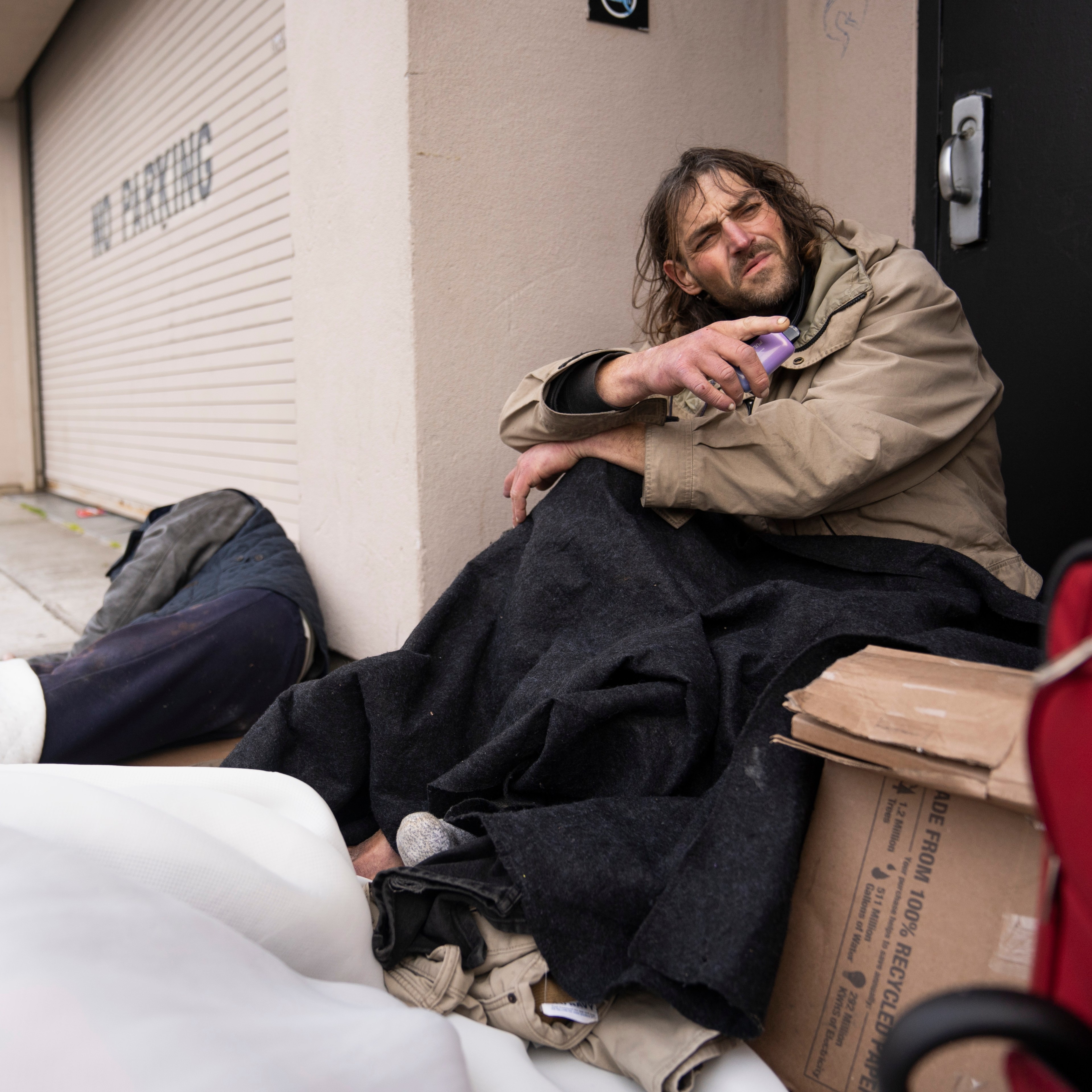 A man sits on the sidewalk wrapped in a blanket, holding an object, beside a closed garage door. Nearby, there's a red suitcase and cardboard boxes.