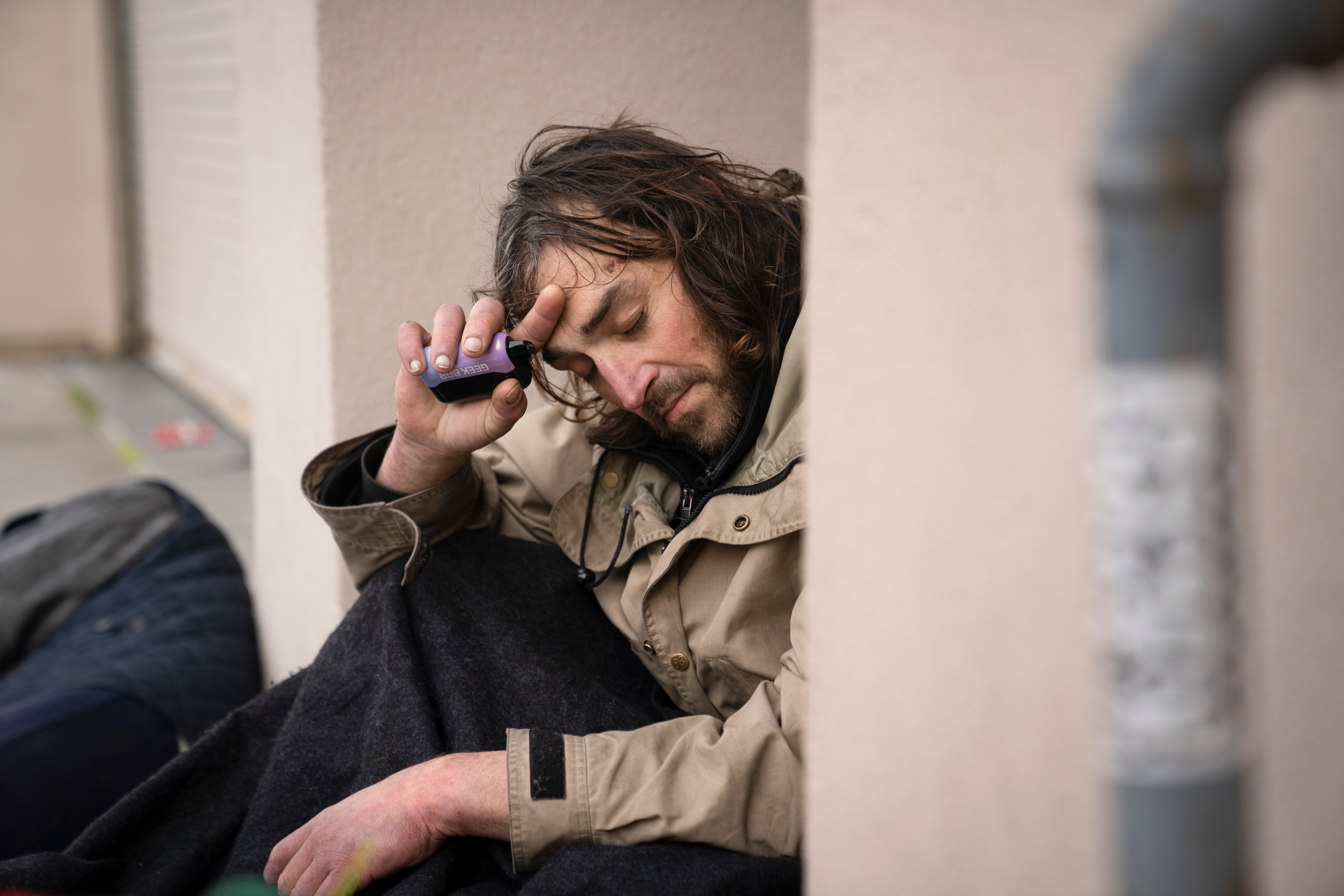 A man with long hair, wearing a beige jacket, sits against a wall, holding his head with one hand and a small bottle with the other, eyes closed.