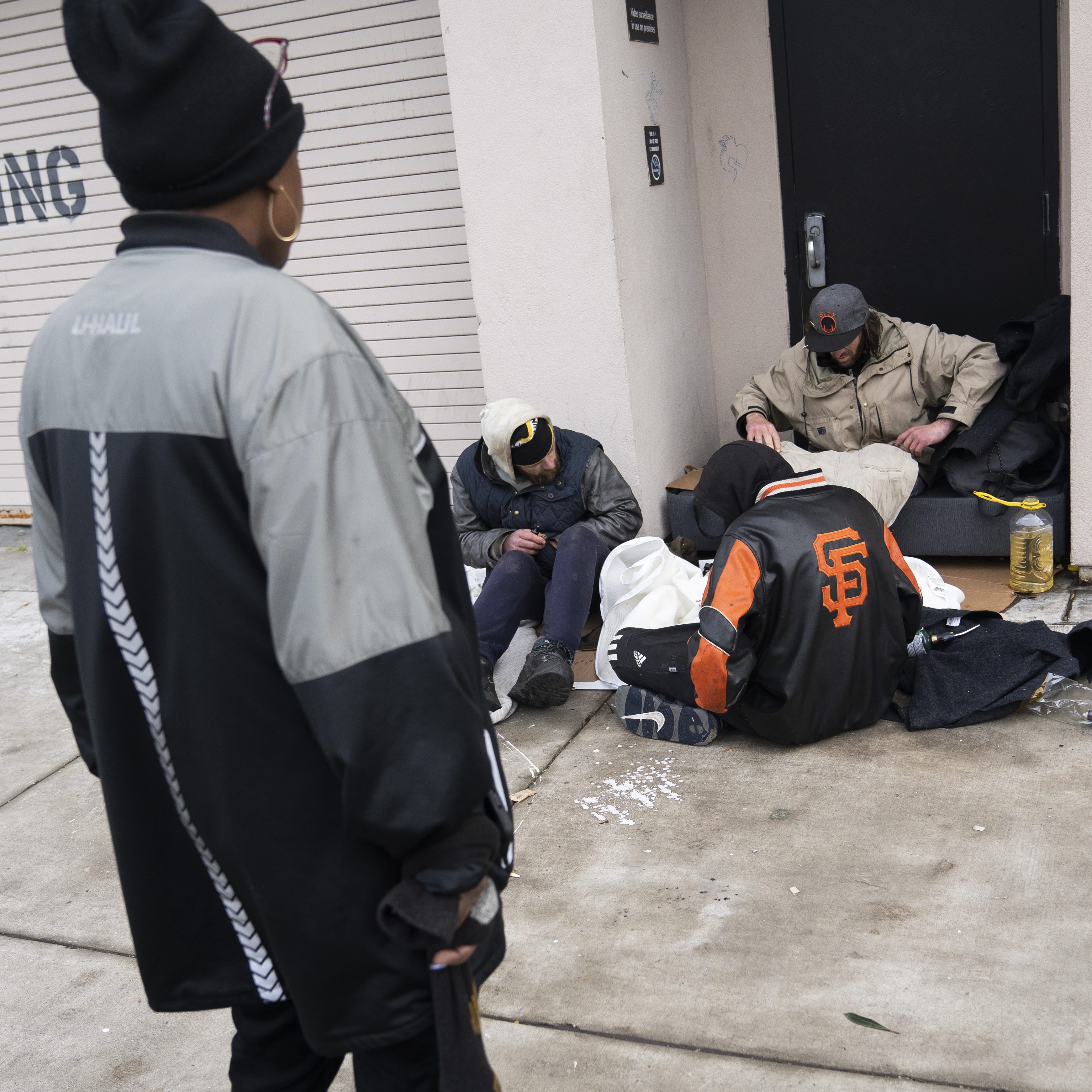 A group of four people sit outside a building entrance on a sidewalk. They wear jackets and beanies, with one person standing and the others sitting or crouching.