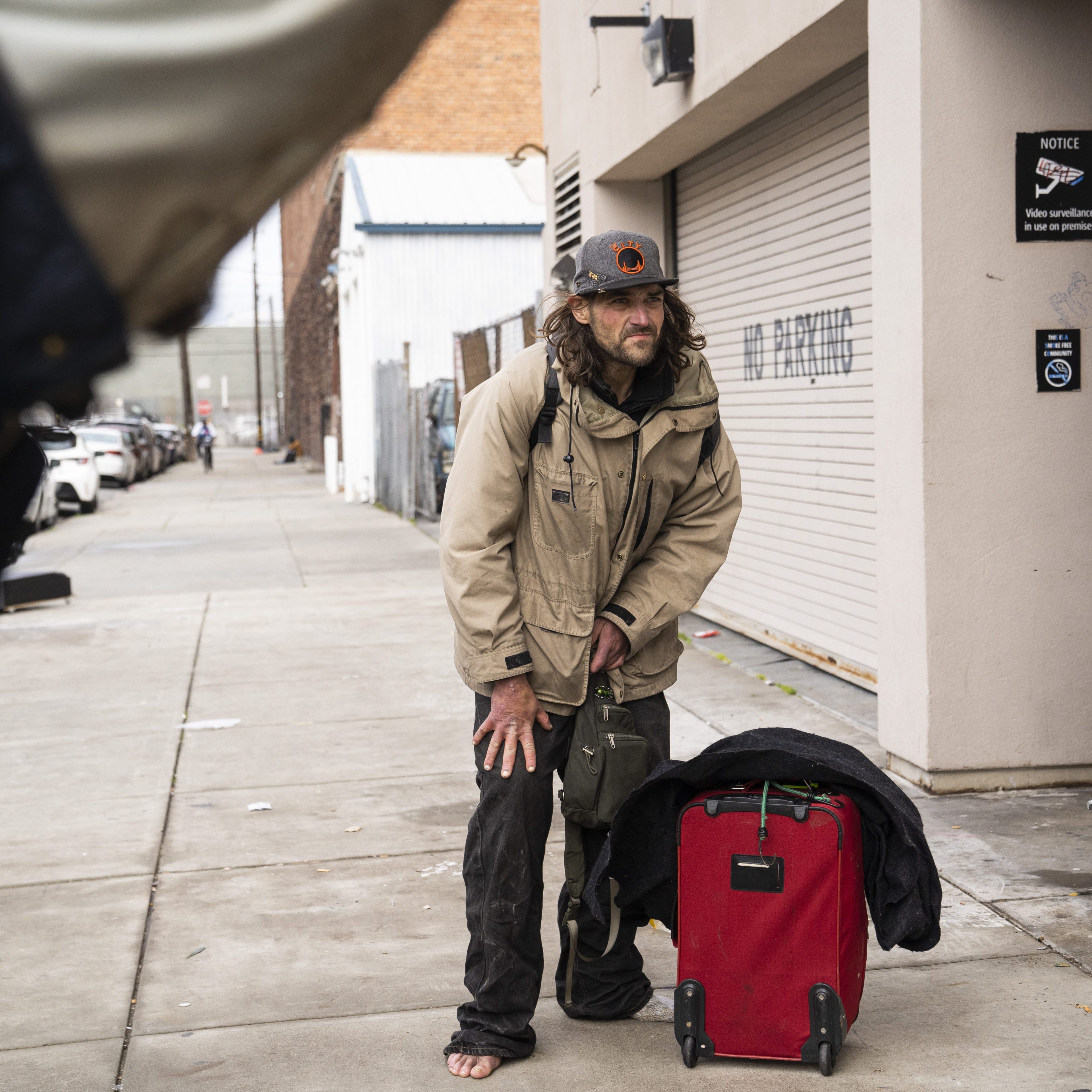 A man in a beige jacket and dark pants stands barefoot on a sidewalk next to a red suitcase. He leans slightly with a determined expression.