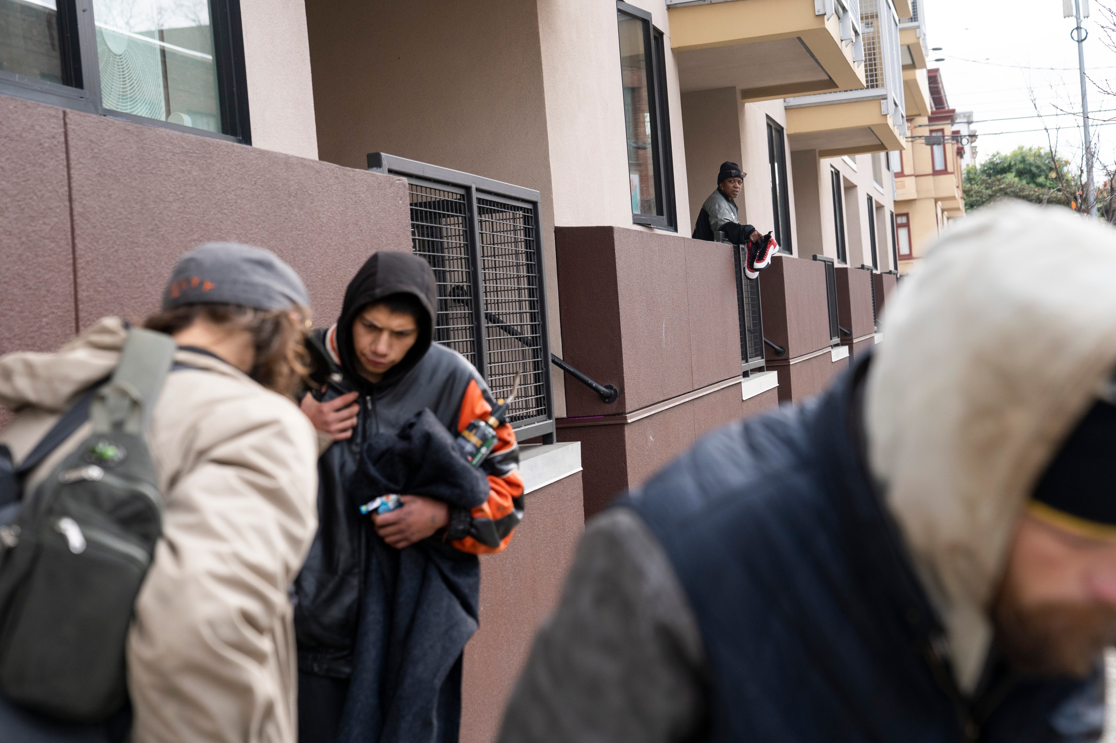 Three people in jackets walk by a building with maroon walls and windows. Another person is leaning on a balcony, holding shoes. The scene feels urban.