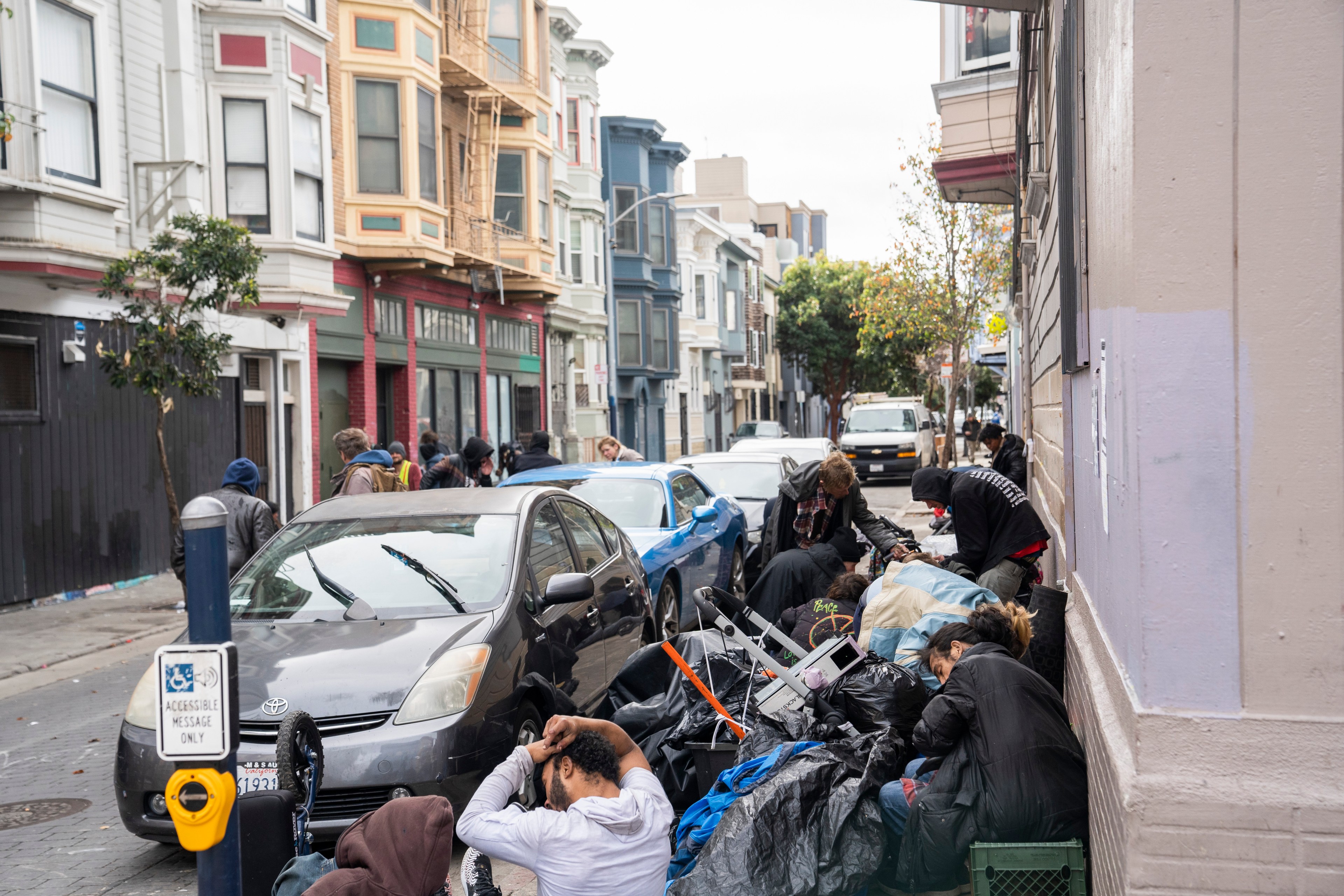 People are gathered on a street cluttered with bags and belongings, beside parked cars and colorful buildings, depicting an urban scene.