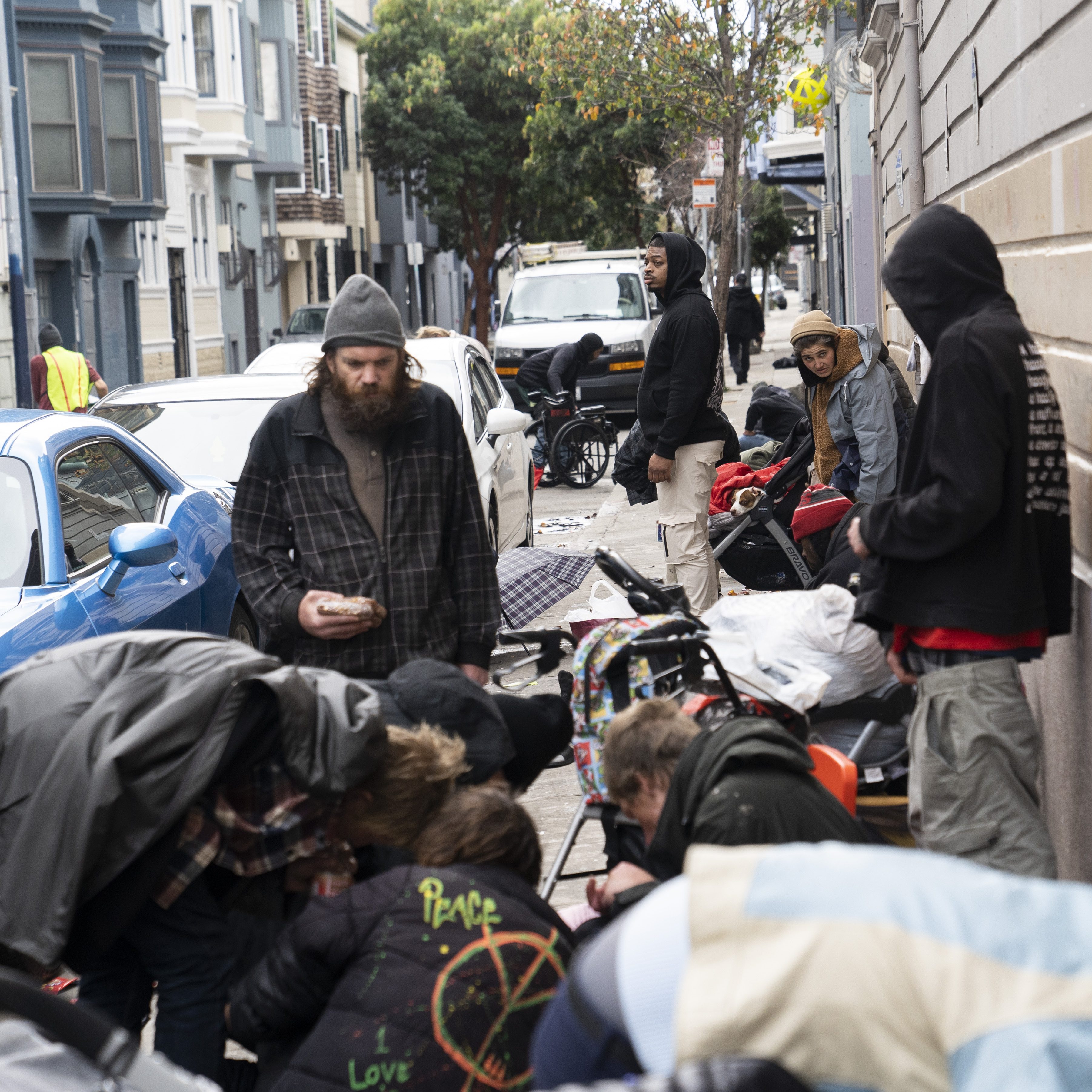 People stand and sit on a sidewalk cluttered with belongings and strollers, beside parked cars and buildings, creating a crowded and busy street scene.