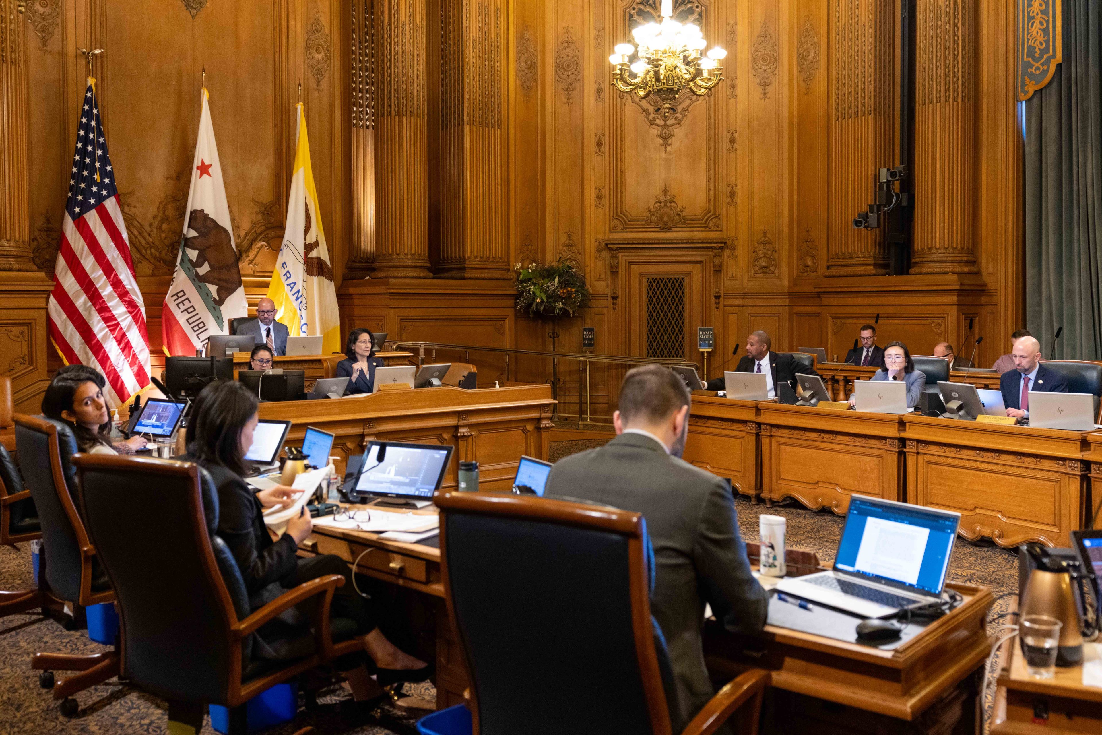 A view of the ornate wooden chamber of the Board of Supervisors with several of them seated at a U shaped table.