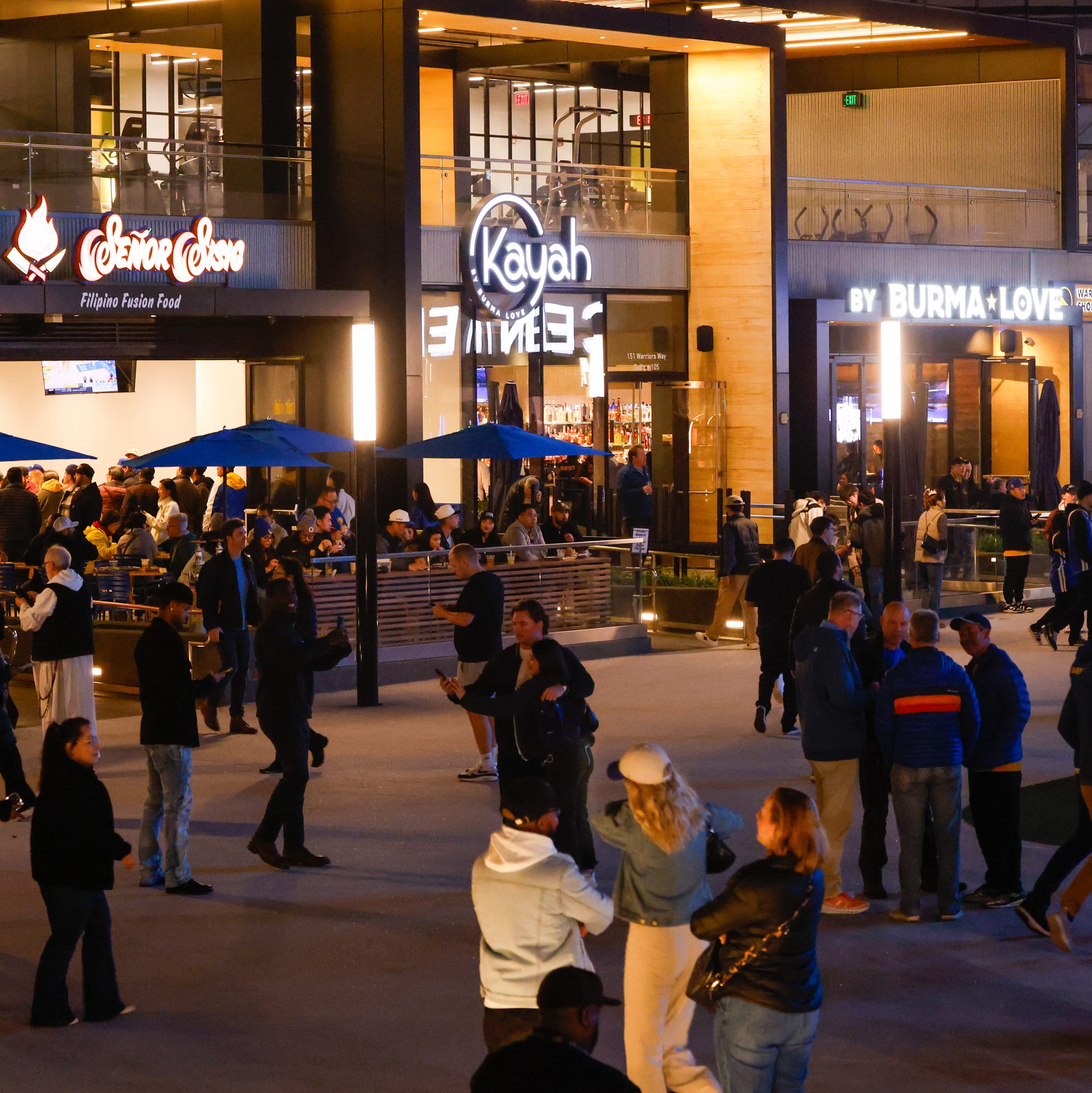 A lively outdoor scene with people socializing near restaurants. Crowds gather under blue umbrellas, enjoying the evening atmosphere and illuminated storefronts.