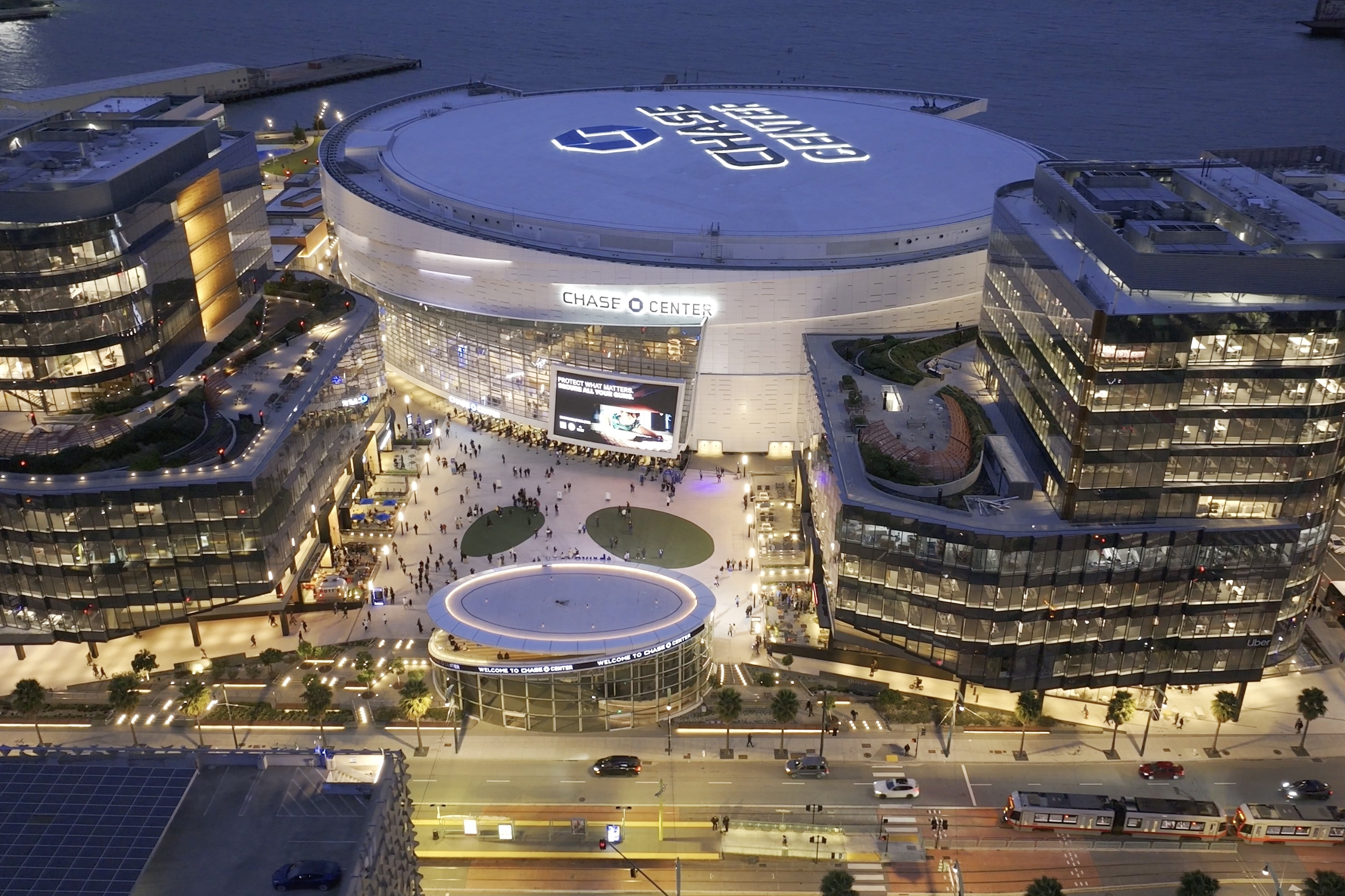 An aerial view of a modern arena illuminated at night, surrounded by bustling crowds and nearby buildings, with waterfront in the background.