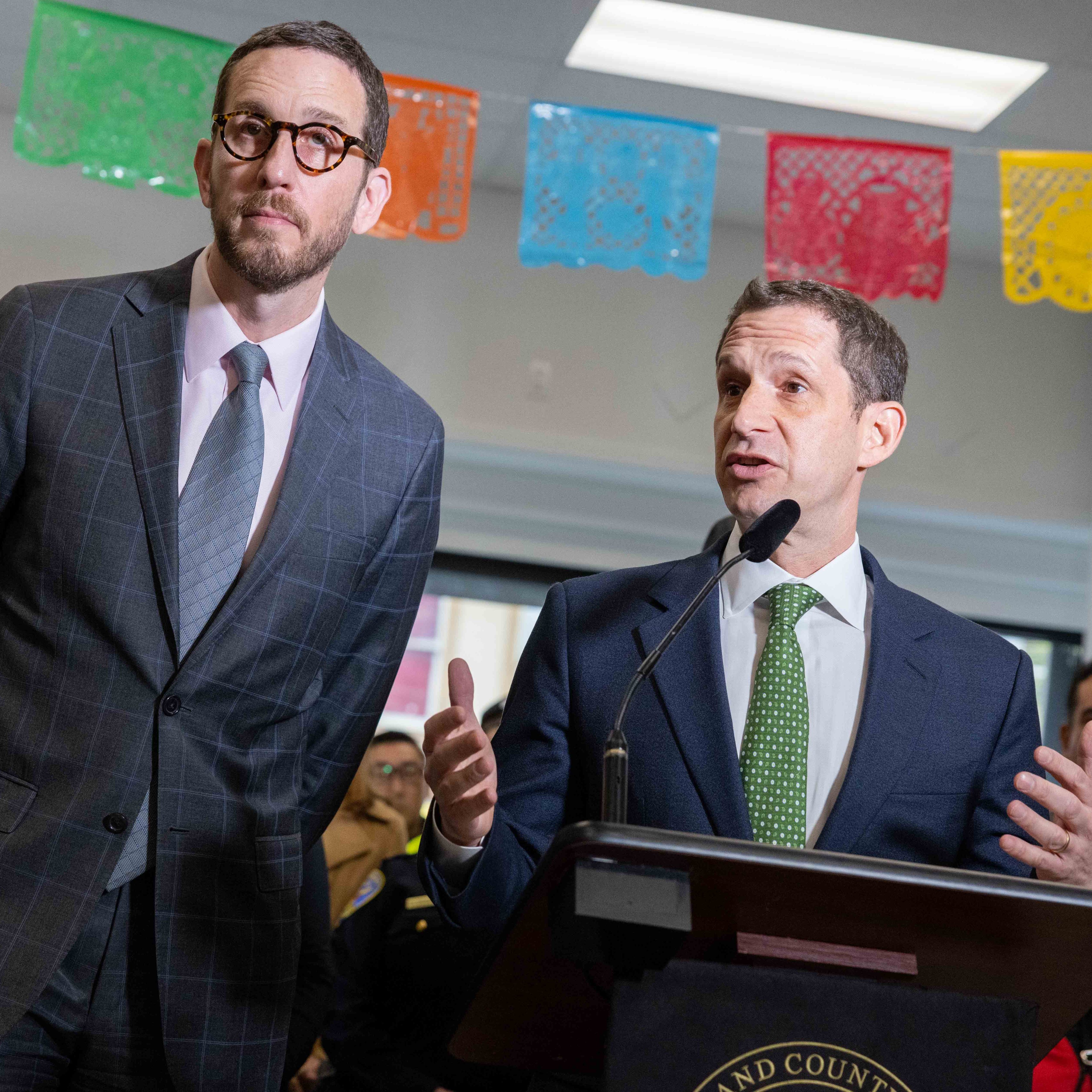 Two men in suits stand at a podium, speaking into a microphone. They are surrounded by people, with colorful paper decorations hanging above.