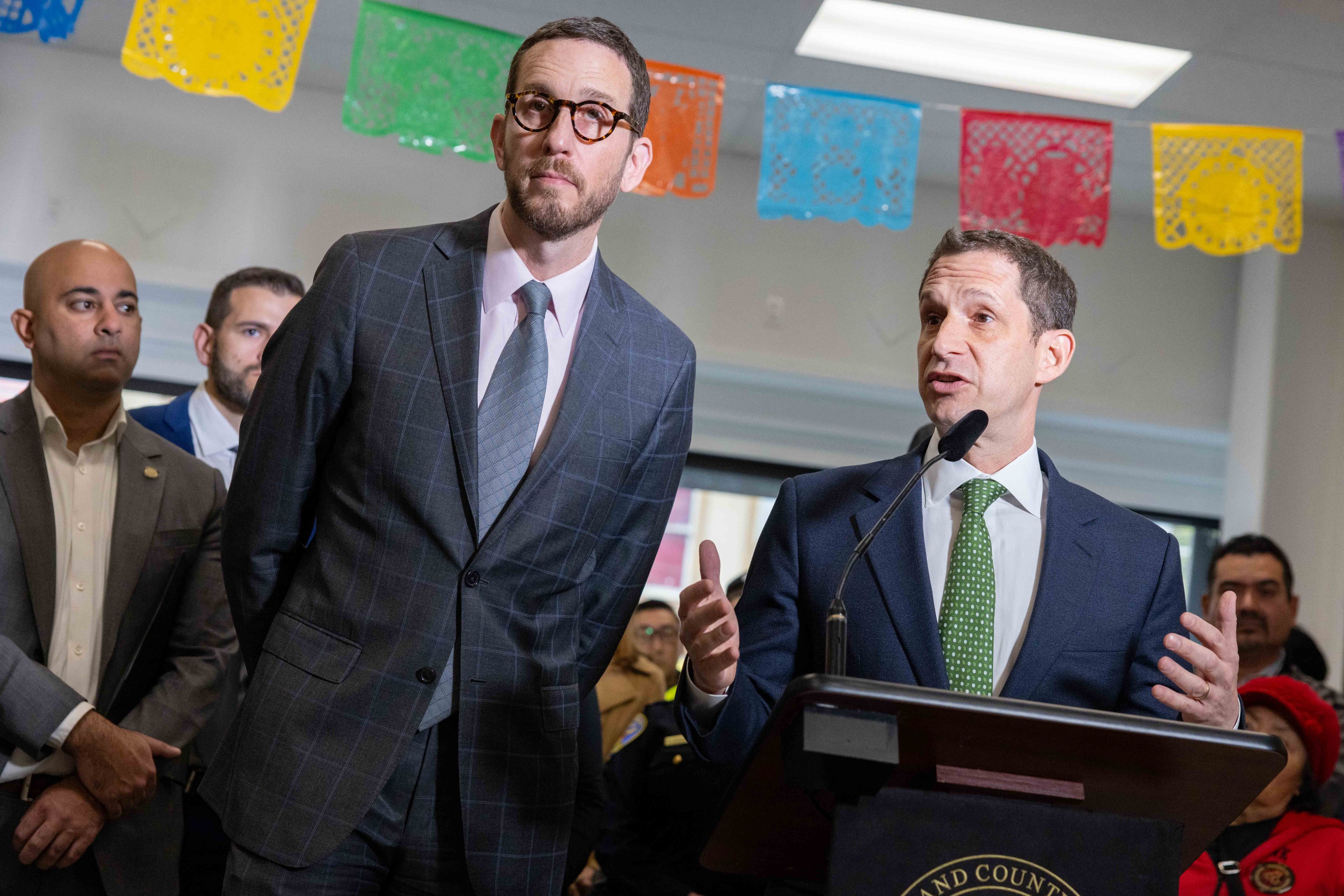 Two men in suits stand at a podium, speaking into a microphone. They are surrounded by people, with colorful paper decorations hanging above.