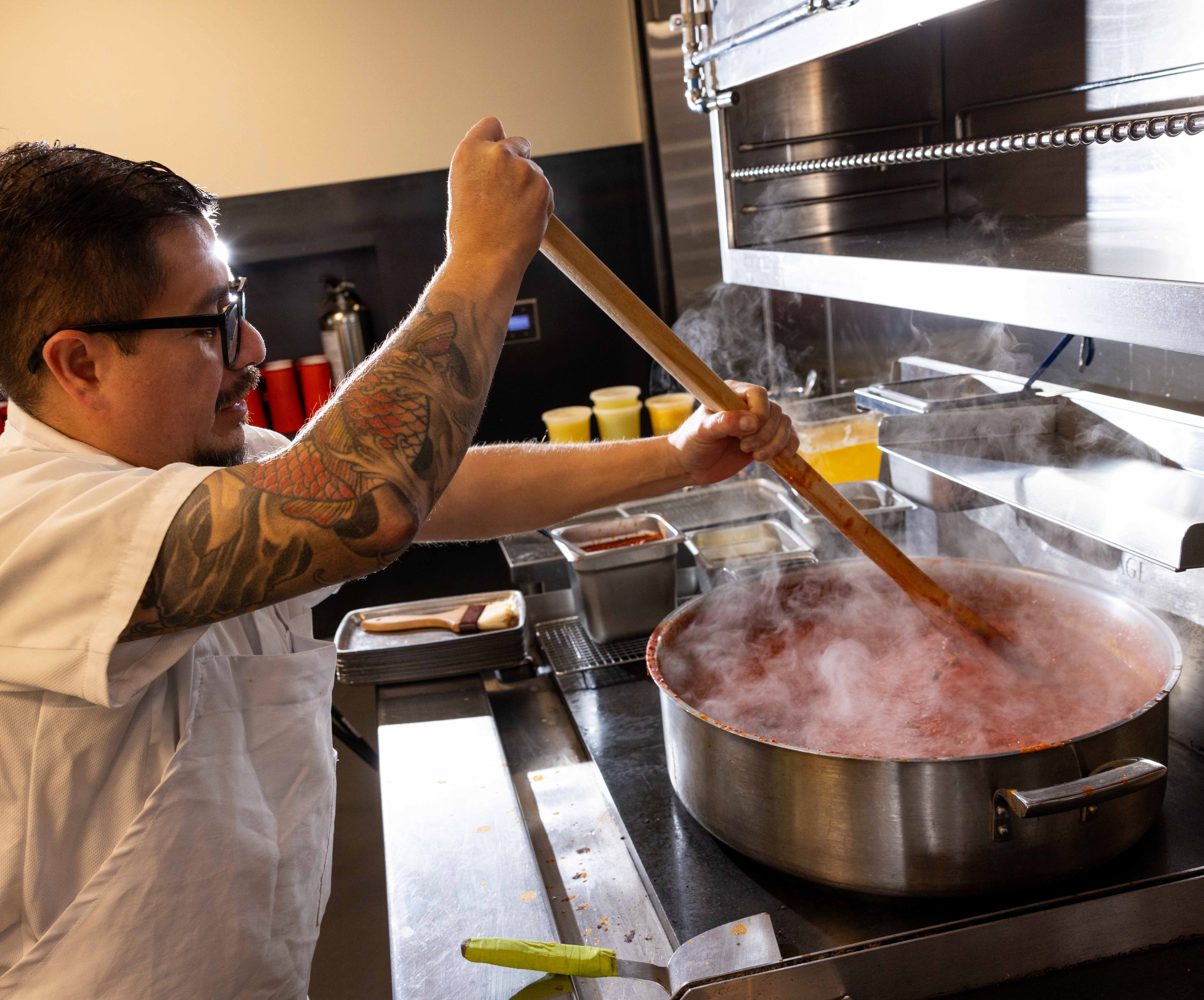 A person with tattoos is stirring a large pot of steaming red sauce in a kitchen. They're using a long wooden spoon. Various kitchen items surround them.