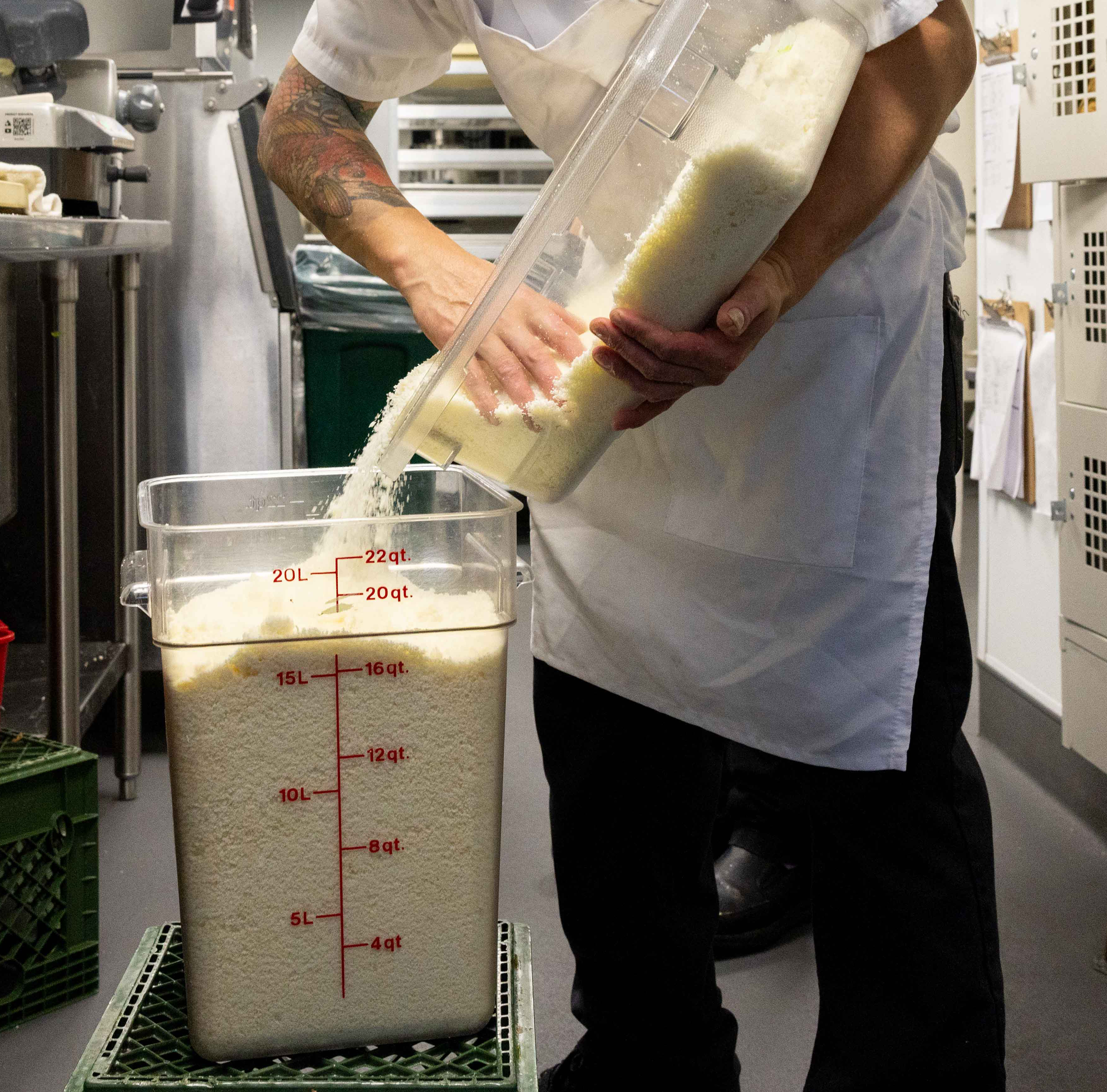 A person in a white apron pours rice from a large container into a bigger, marked container on a green crate in a kitchen setting.