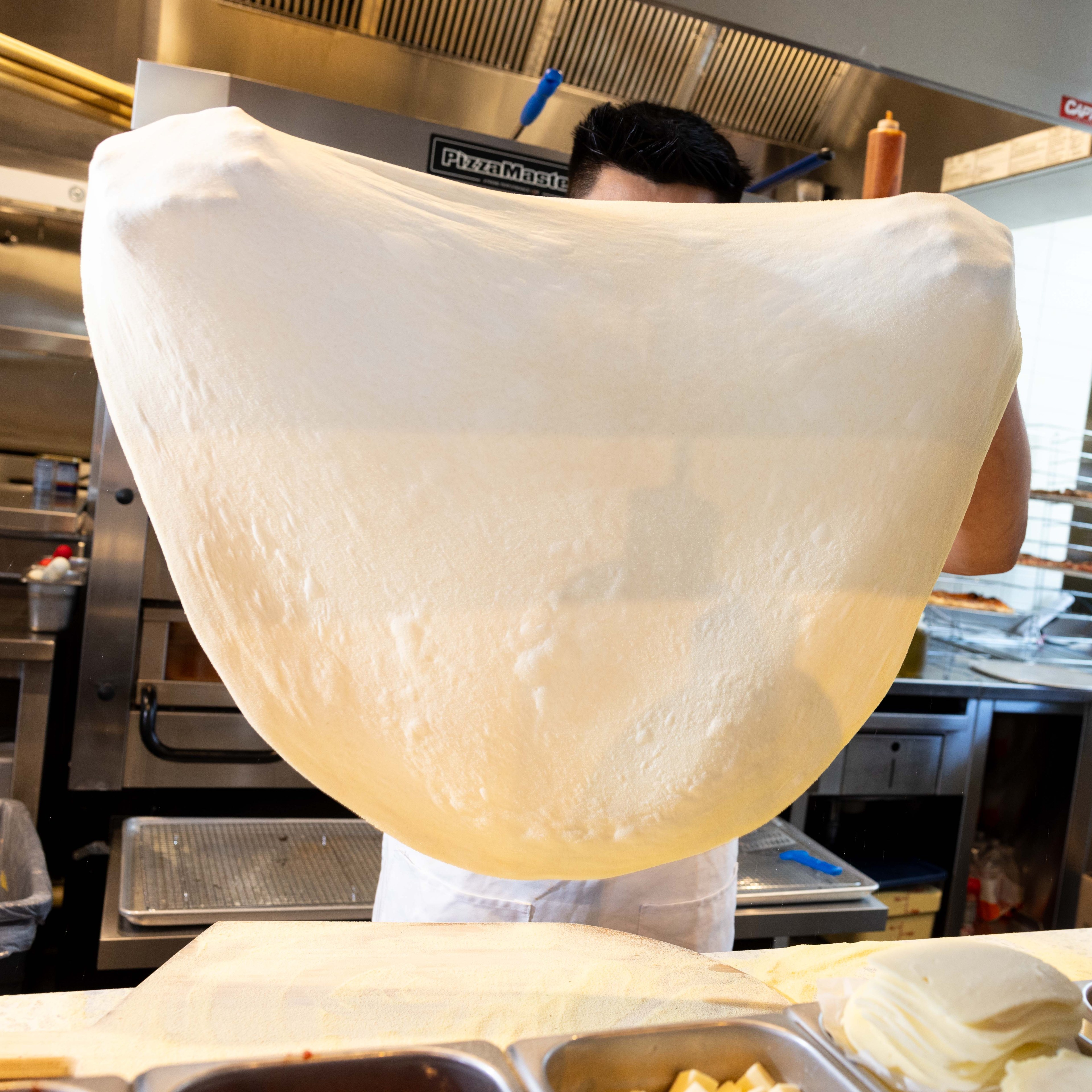 A person in a kitchen stretches a large piece of dough, obscuring their face. The background shows kitchen appliances and countertop containers.