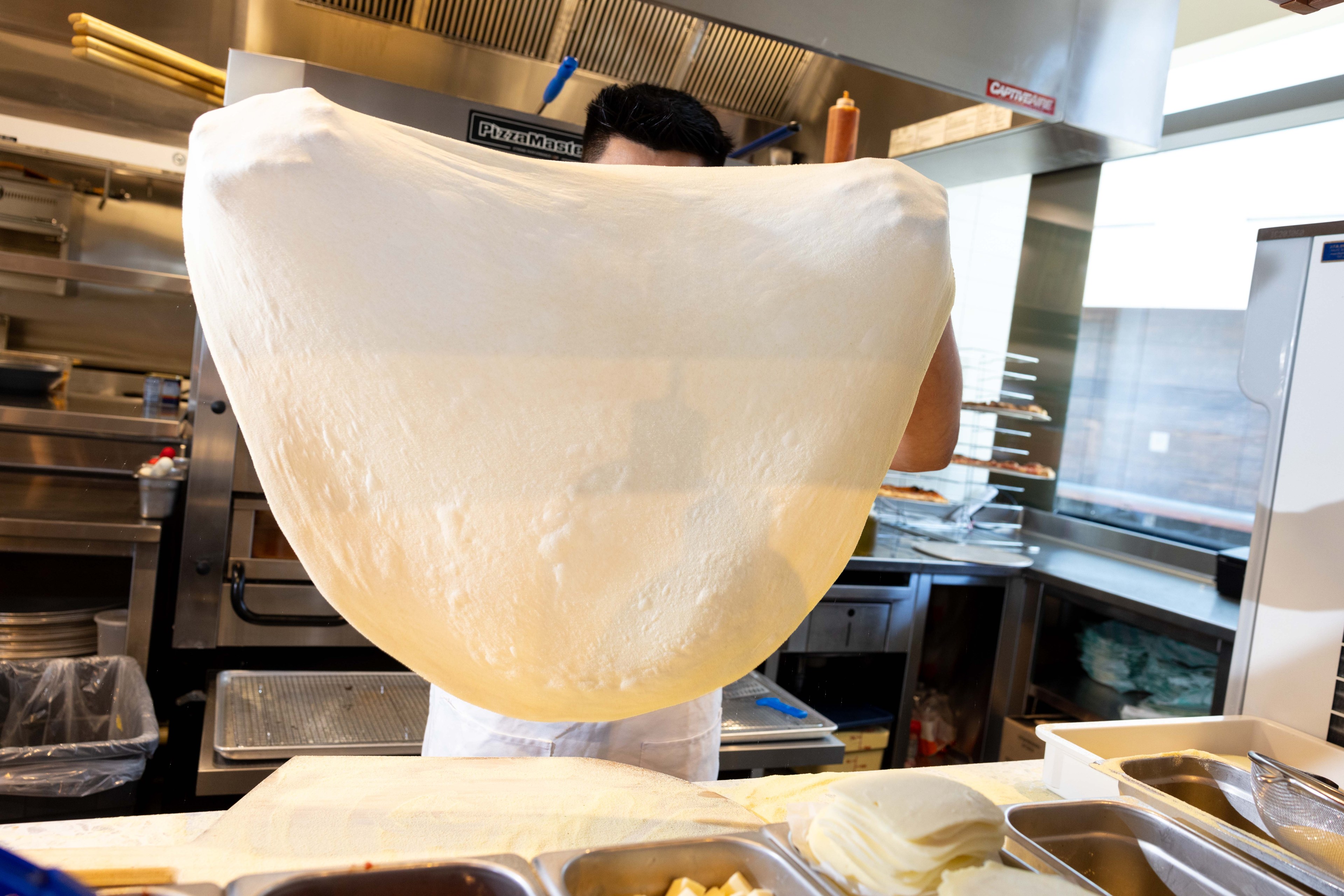 A person in a kitchen stretches a large piece of dough, obscuring their face. The background shows kitchen appliances and countertop containers.