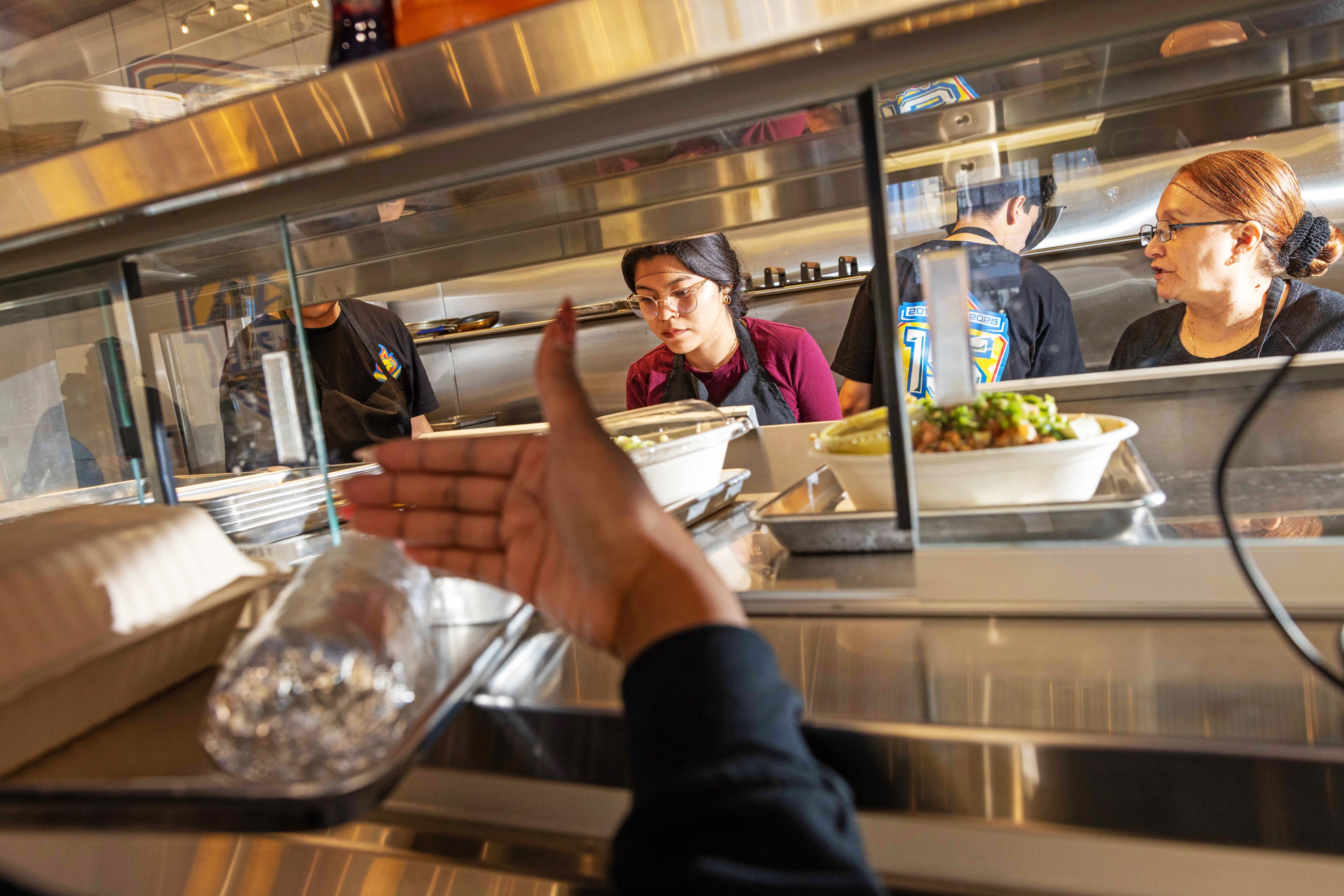 A hand reaches out for a foil-wrapped burrito on a counter. Behind the counter, three people are working, with food bowls and trays in view.