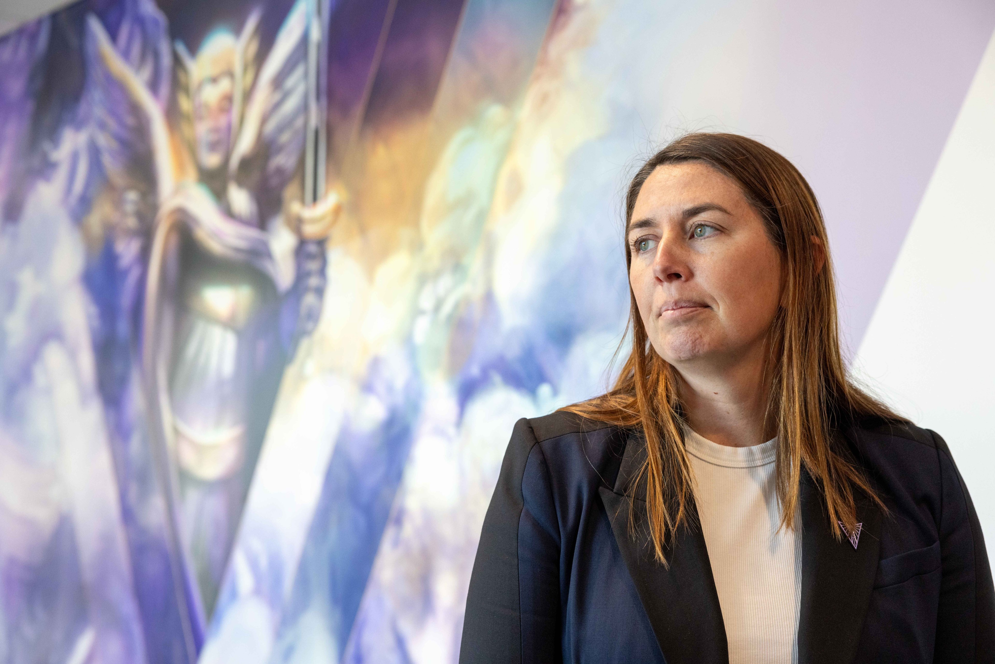 A woman with long hair, wearing a blazer and white top, stands against a colorful mural of an angelic figure holding a sword.