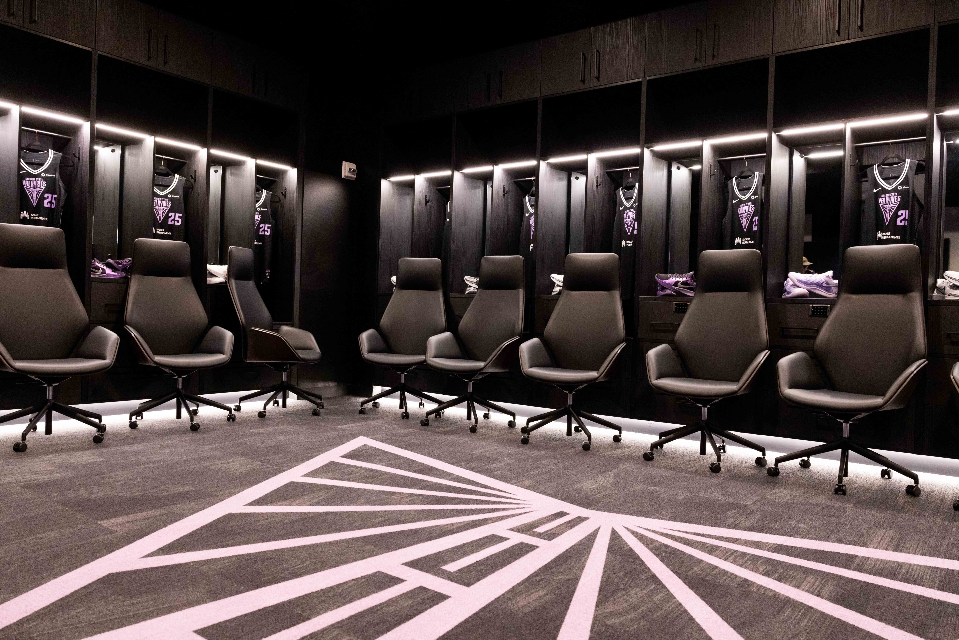 A sleek locker room with black chairs on wheels, illuminated lockers holding black and purple sports jerseys, sneakers, and a patterned carpet.