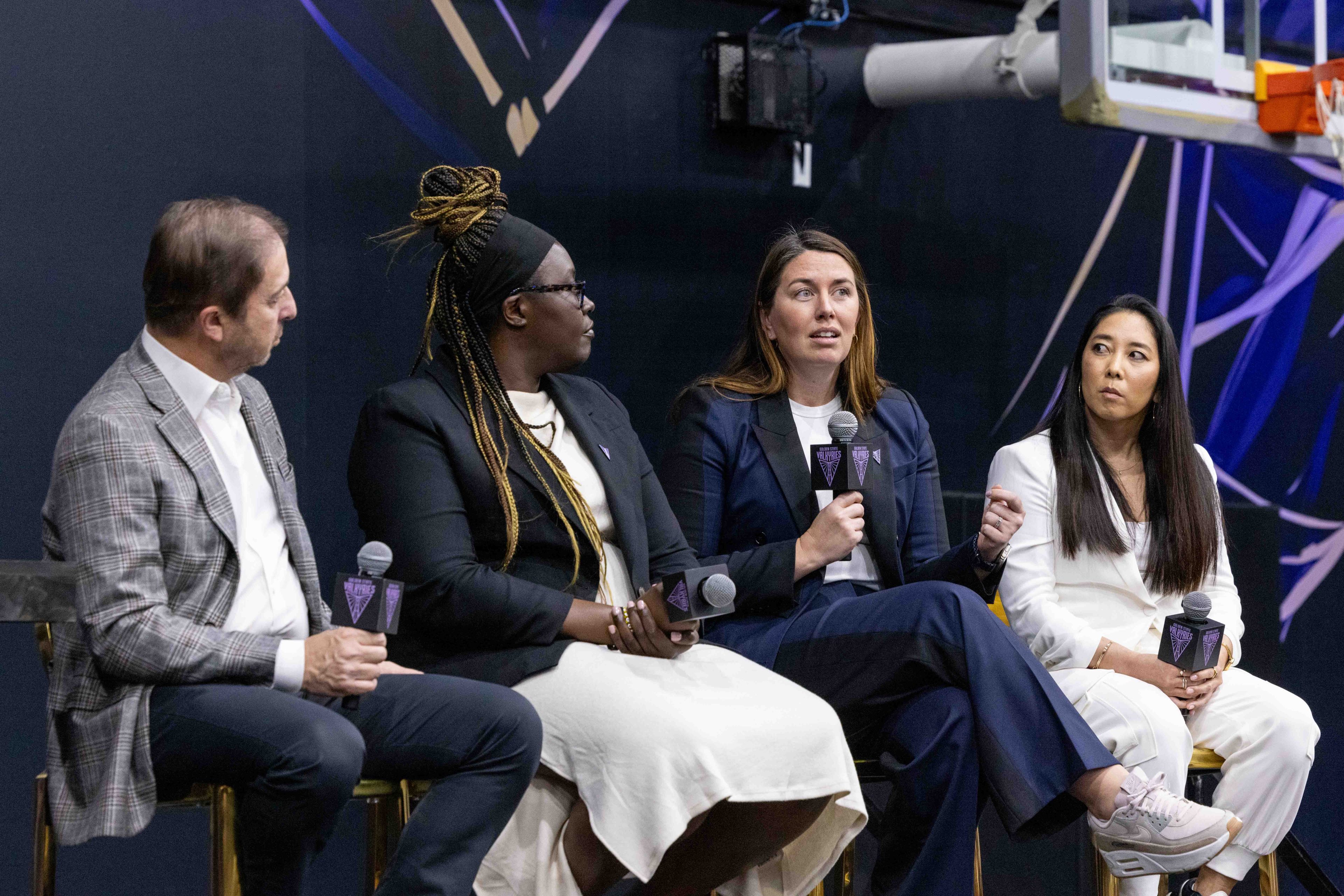 Four individuals are seated on a panel, each holding a microphone. They're engaged in a discussion in front of a background featuring a basketball hoop.