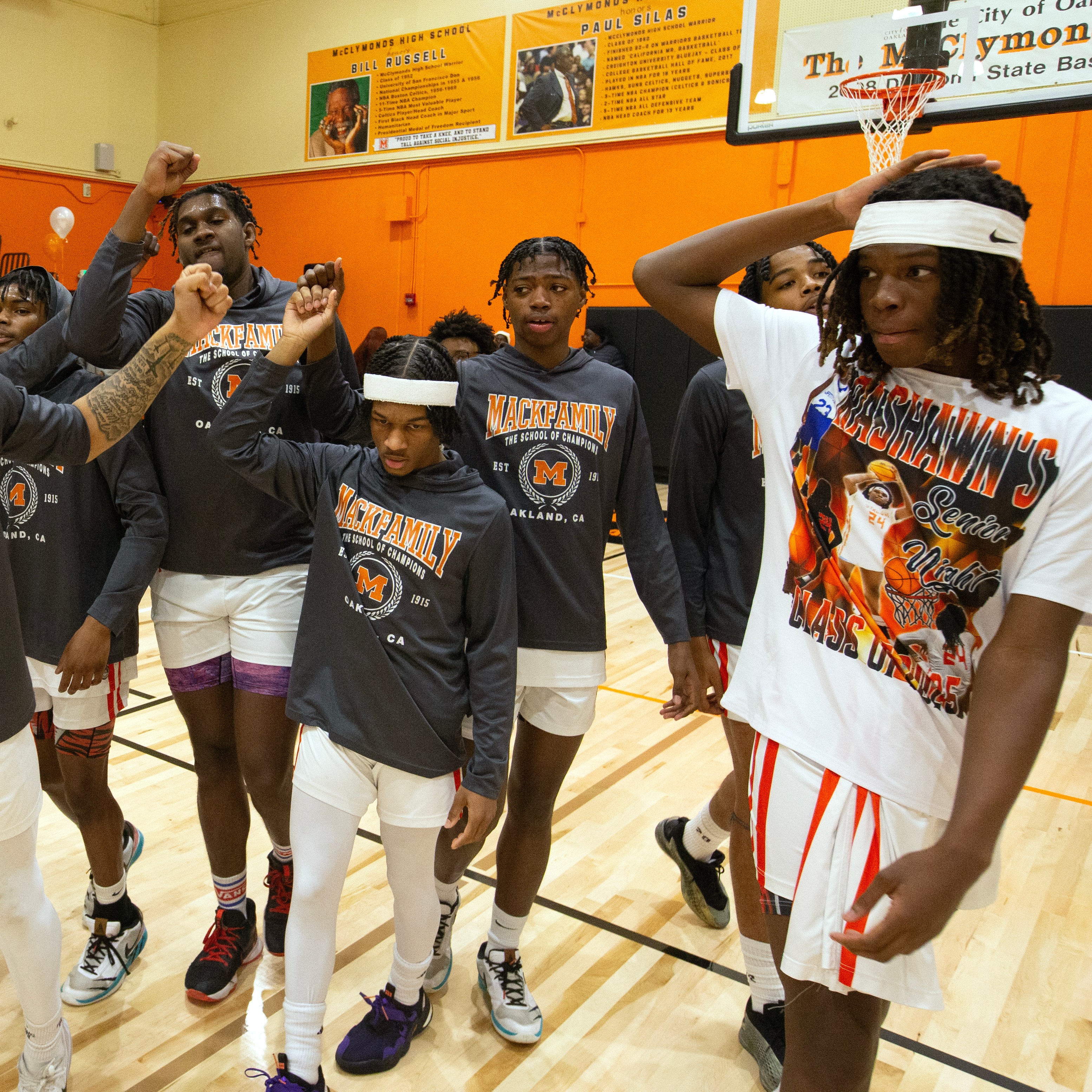 A group of basketball players in matching "Mack Family" shirts are on a court, some with headbands, appearing enthusiastic and energetic.