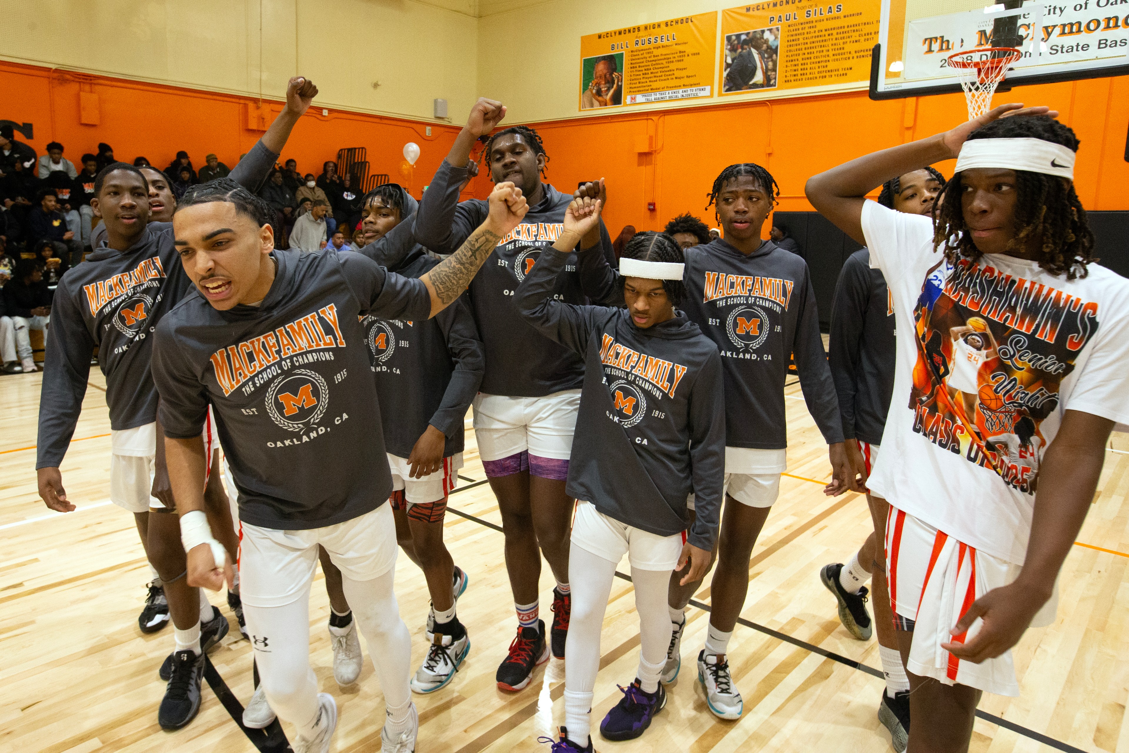 A group of basketball players in matching "Mack Family" shirts are on a court, some with headbands, appearing enthusiastic and energetic.