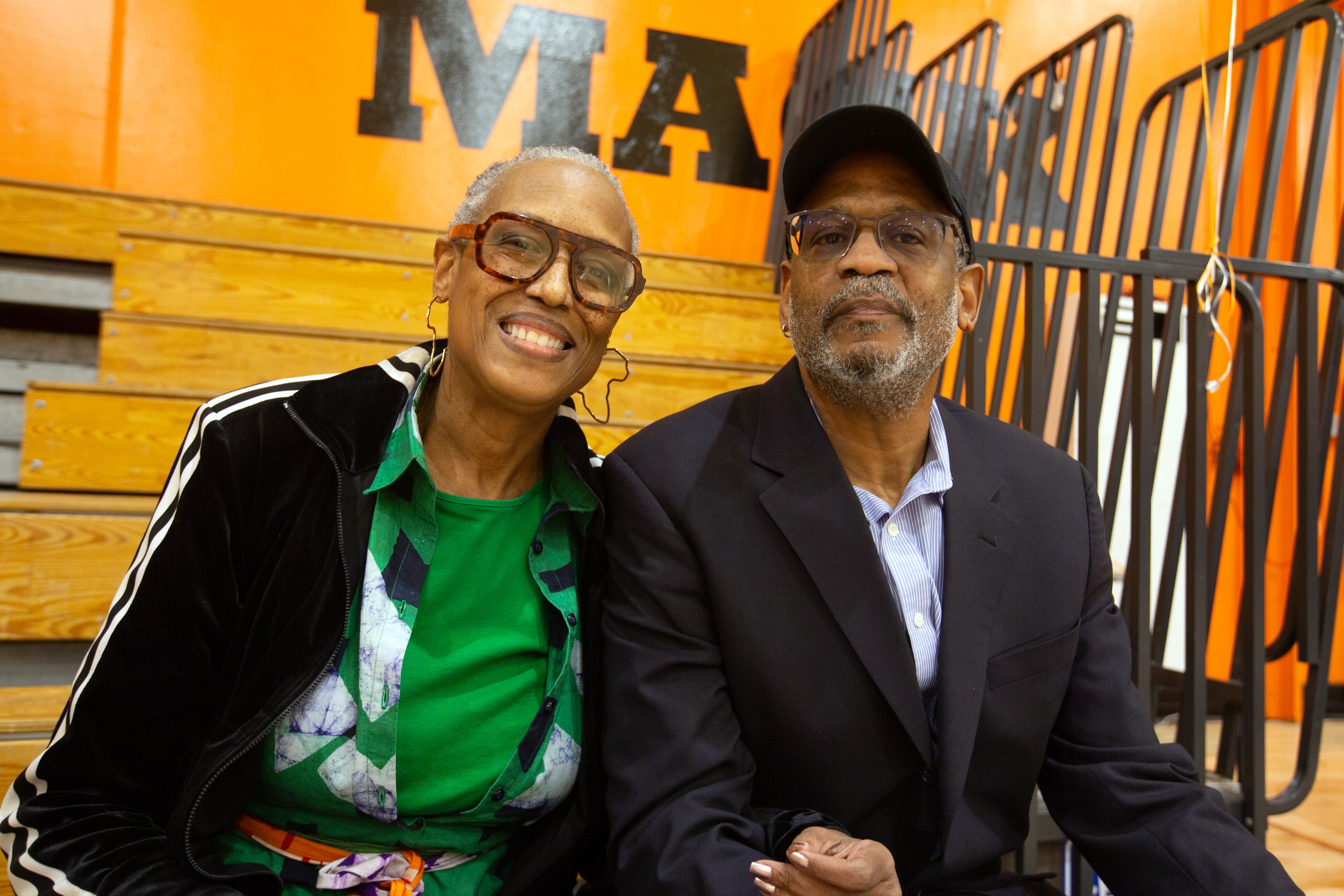 An elderly couple is sitting together, smiling. The woman wears glasses and a green shirt, while the man is in a suit and cap. They're indoors, on bleachers.