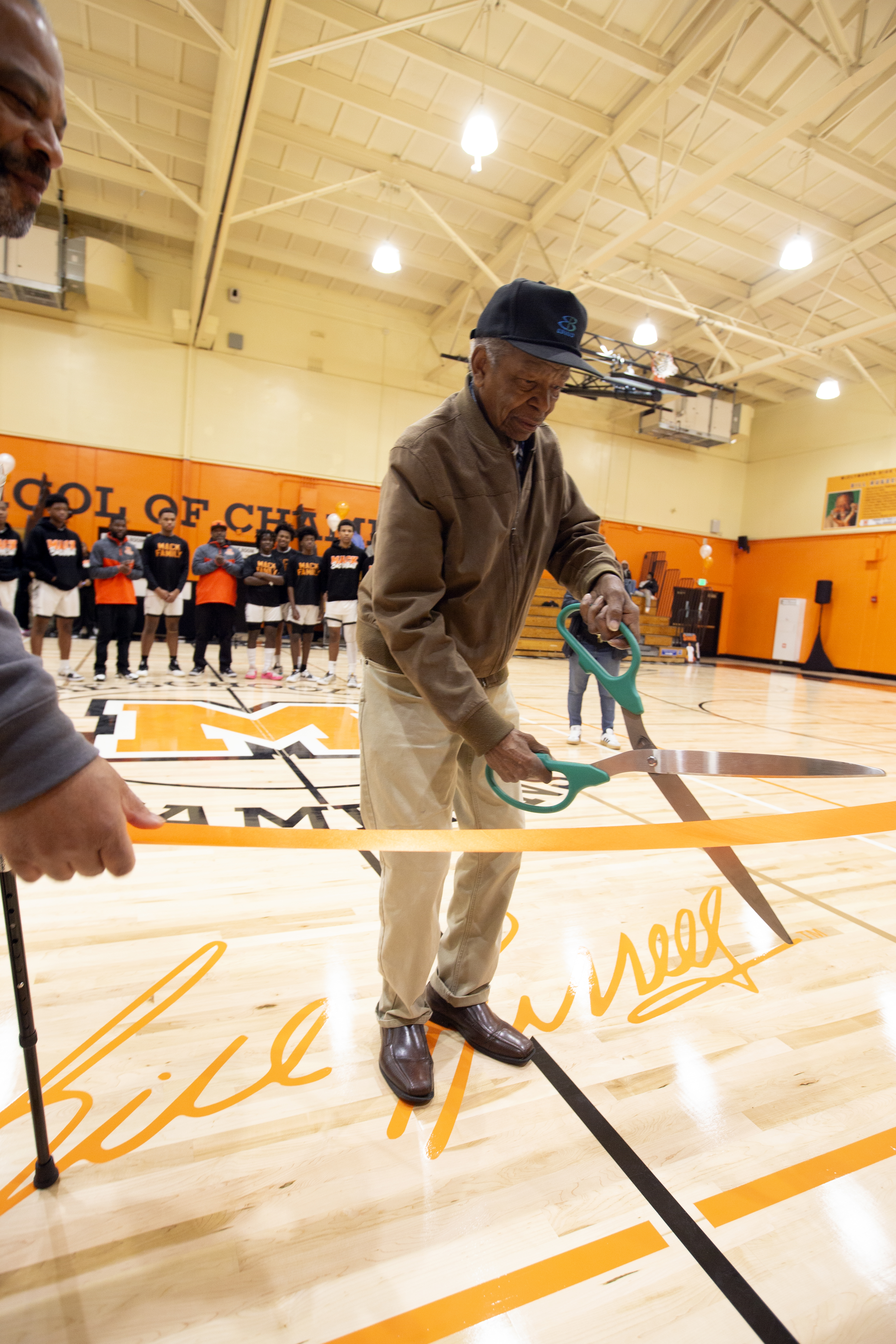 An older man is cutting a ribbon with oversized scissors on a basketball court. People are standing in the background, wearing matching outfits.