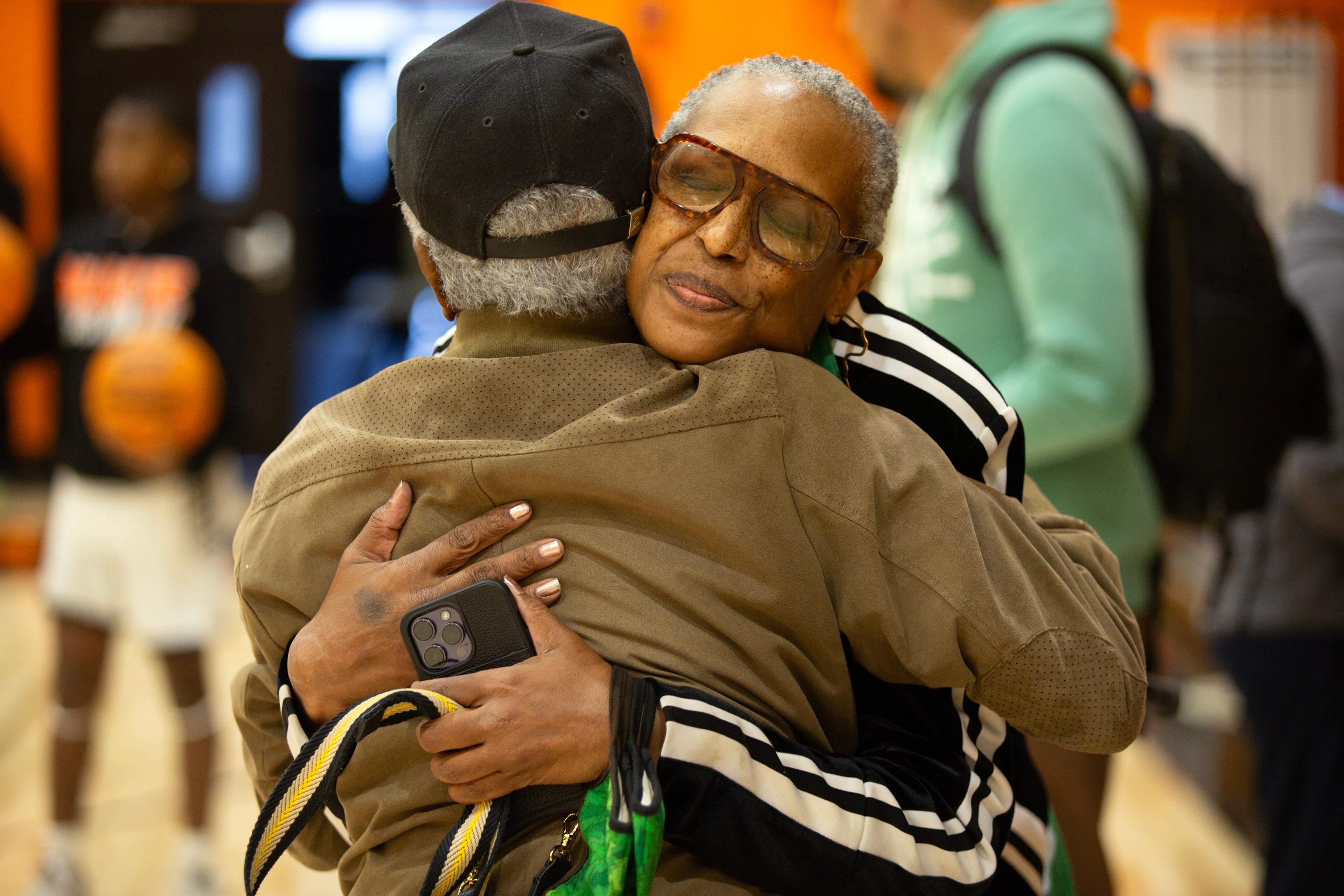 Two people are hugging warmly in a sports setting. One holds a phone, and both appear happy and affectionate. Others are blurred in the background.