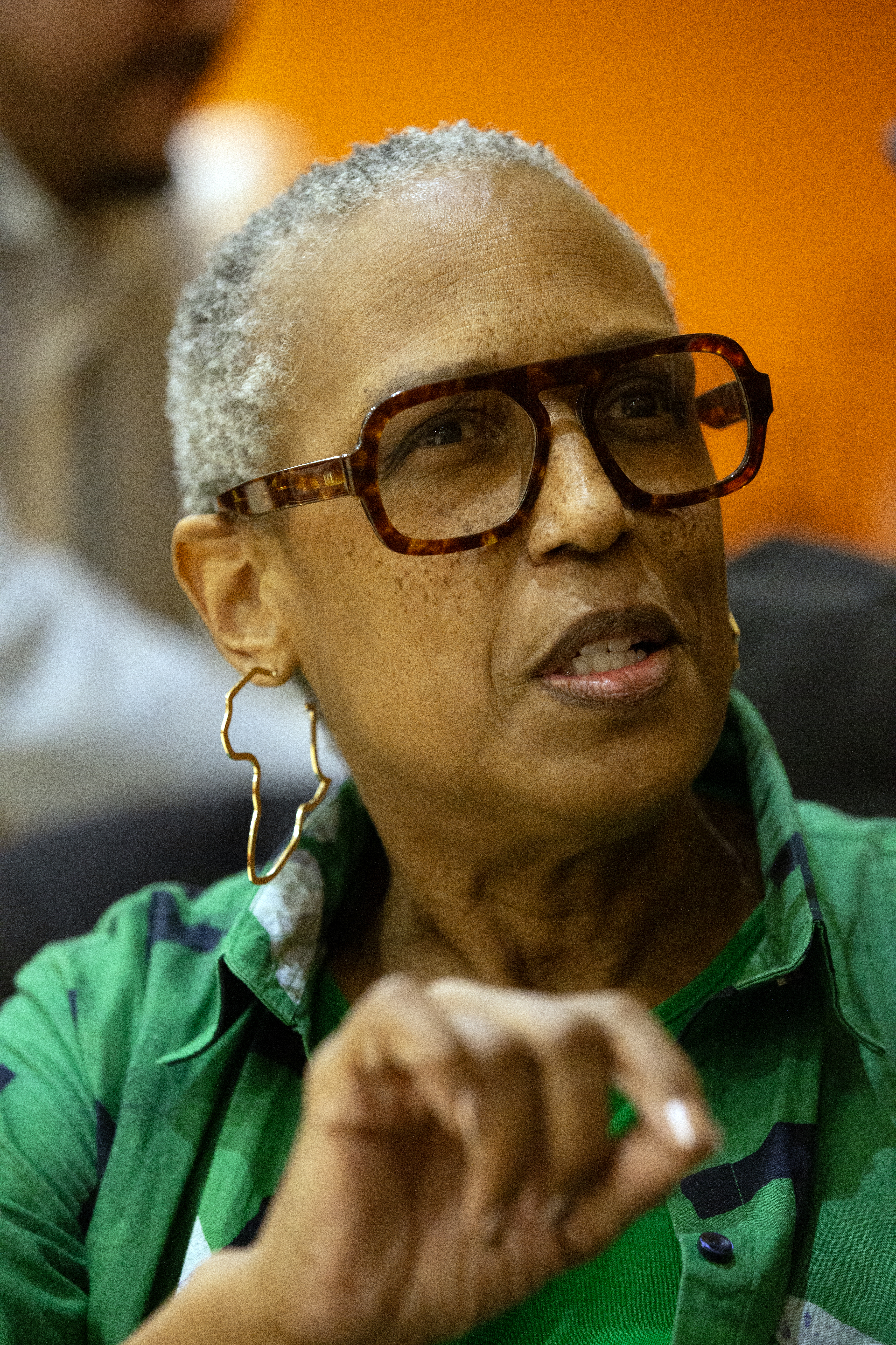 A person with short gray hair and large tortoiseshell glasses is speaking, wearing hoop earrings and a green patterned shirt. An orange background is visible.