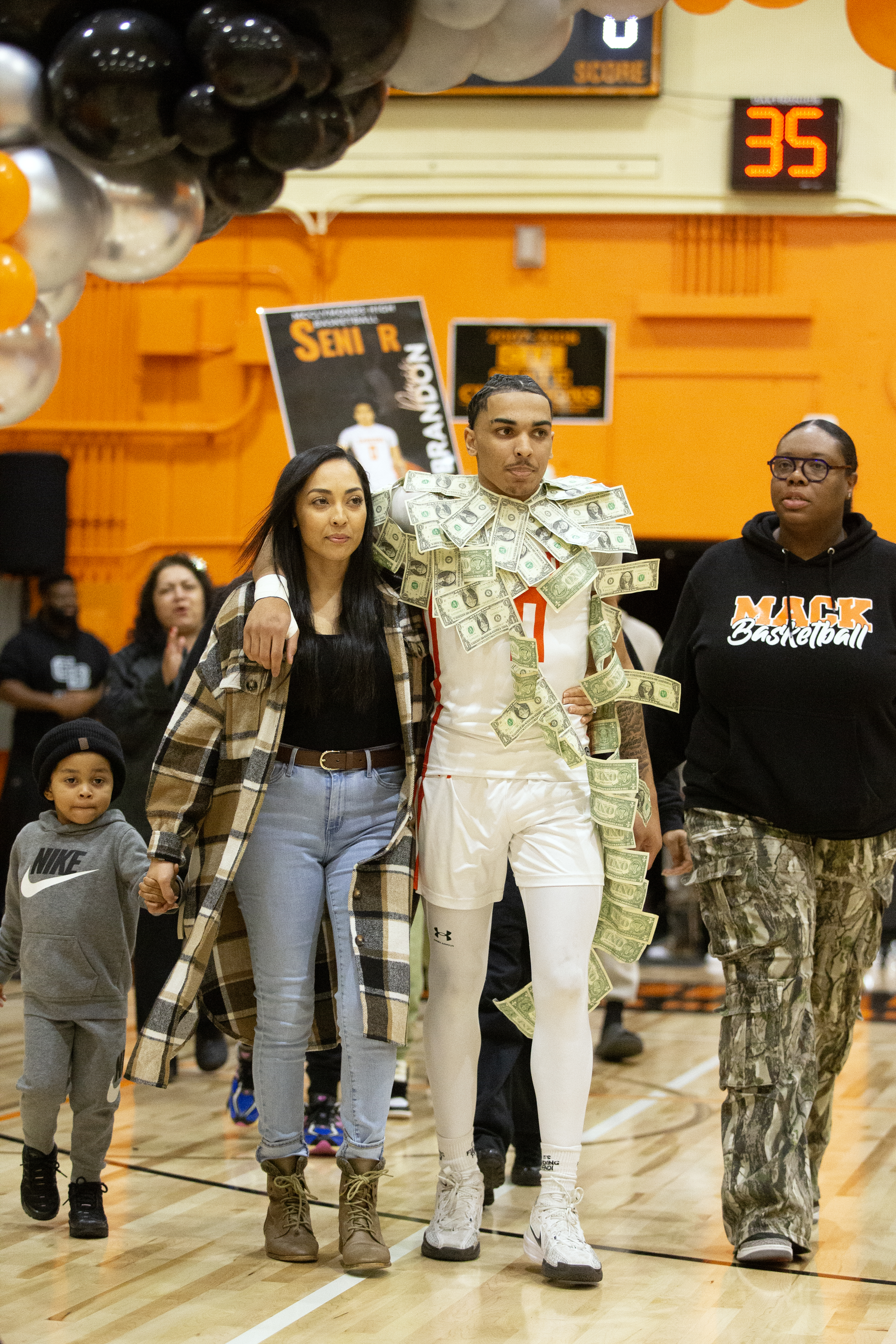 A basketball player, draped in dollar bills, walks arm-in-arm with two women on a gym floor. A child holds hands with one woman. Balloons decorate above.