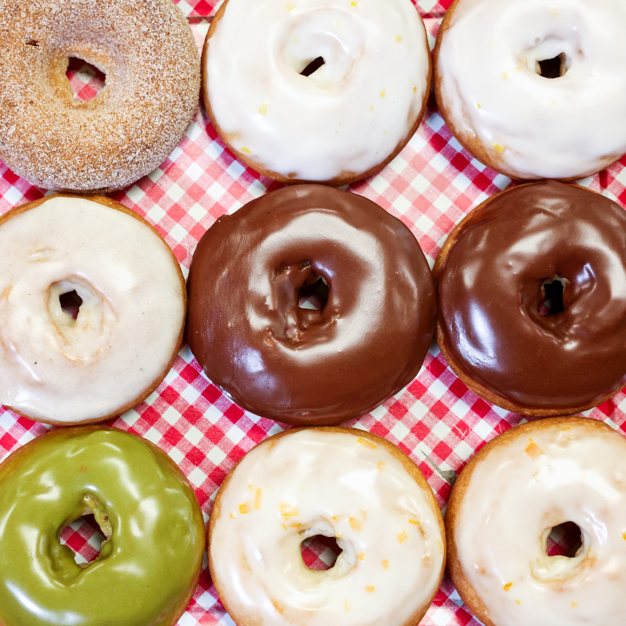 Nine donuts are arranged on a red checkered cloth. They feature different toppings: cinnamon sugar, white glaze, chocolate, and green matcha.