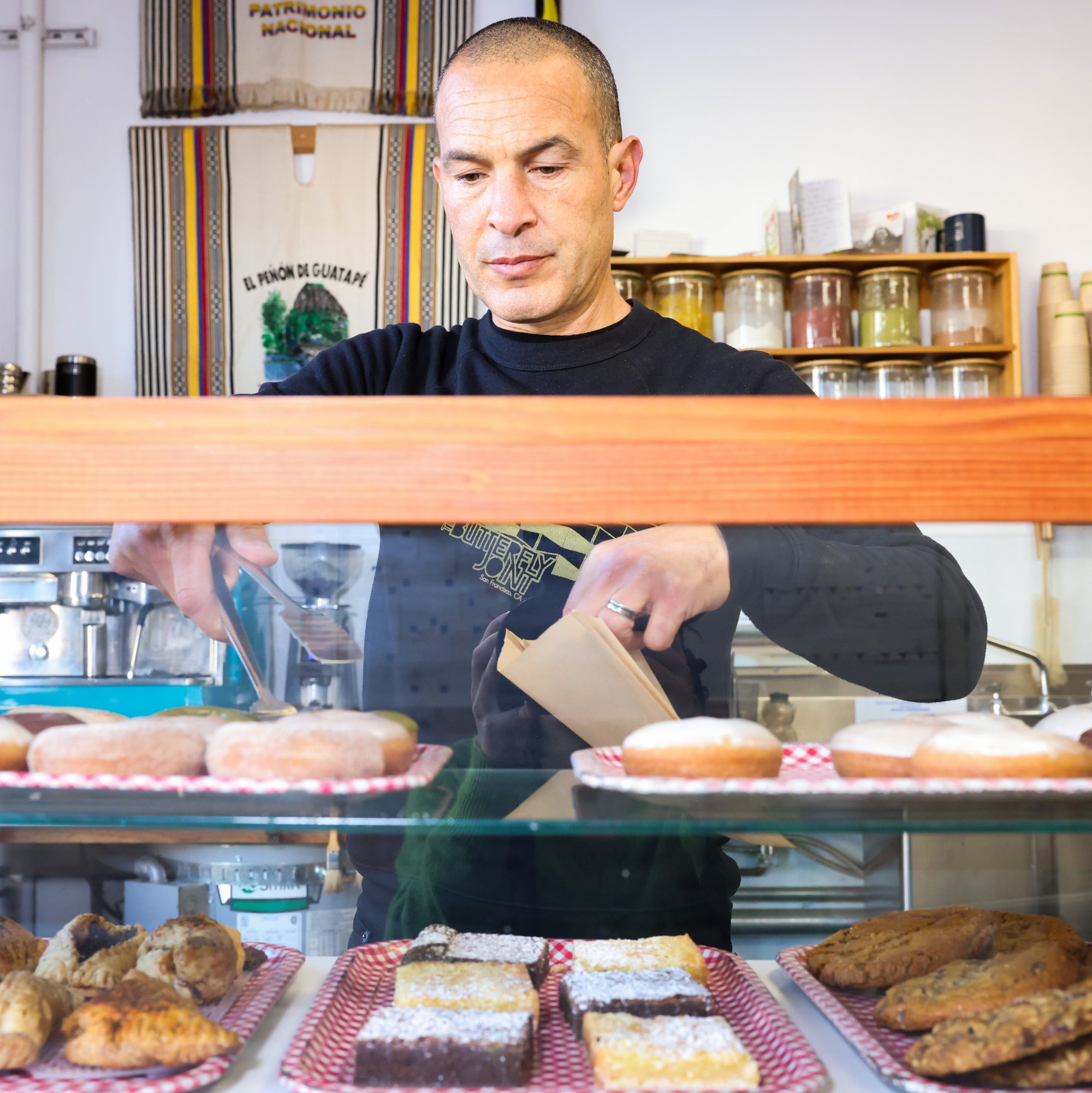 A person stands behind a bakery display case, using tongs to pick donuts. Various pastries are arranged on red-checkered trays, with jars in the background.