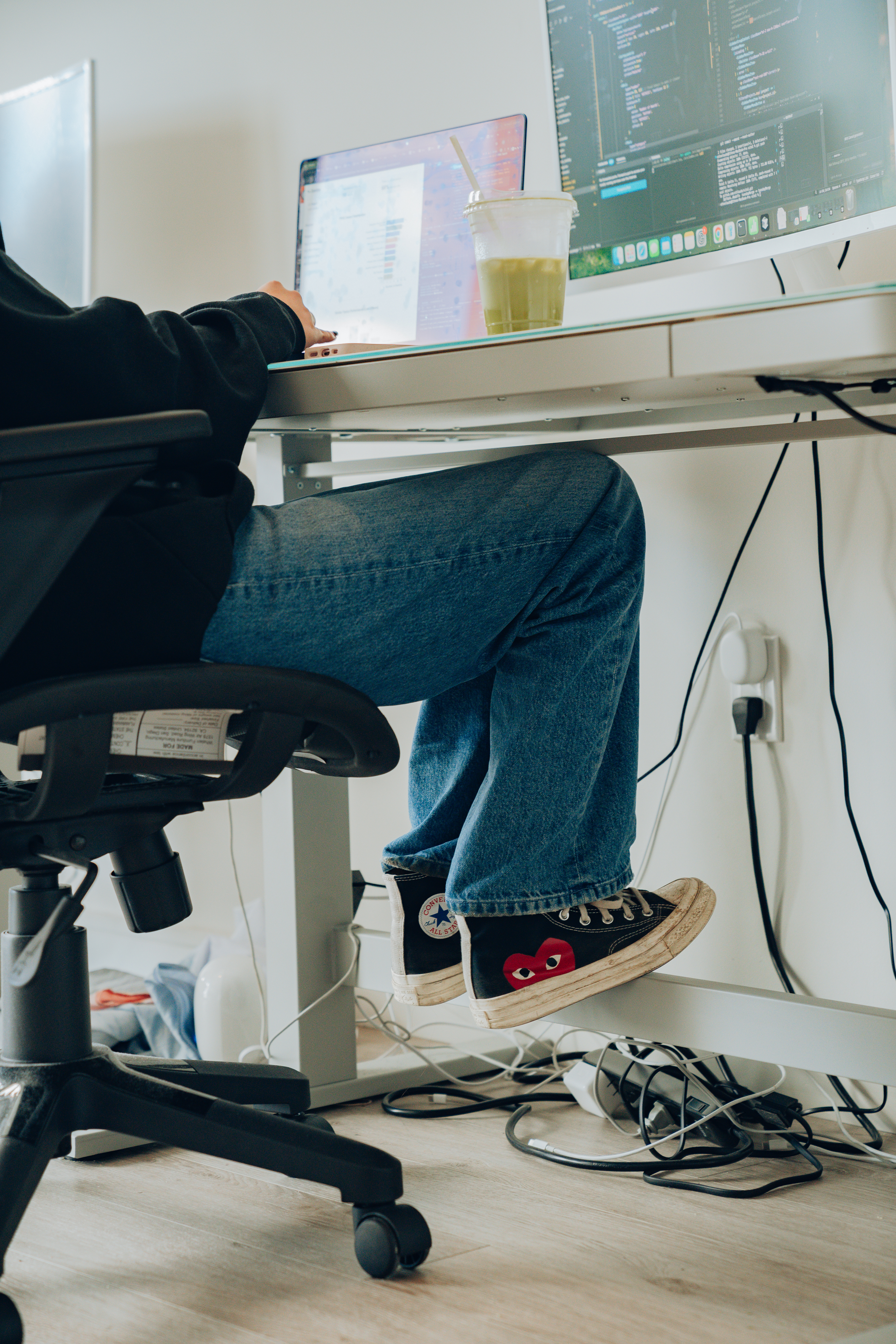 A person sits at a desk using a laptop and monitor with visible code. They wear jeans and sneakers with a distinctive heart design. A drink rests nearby.