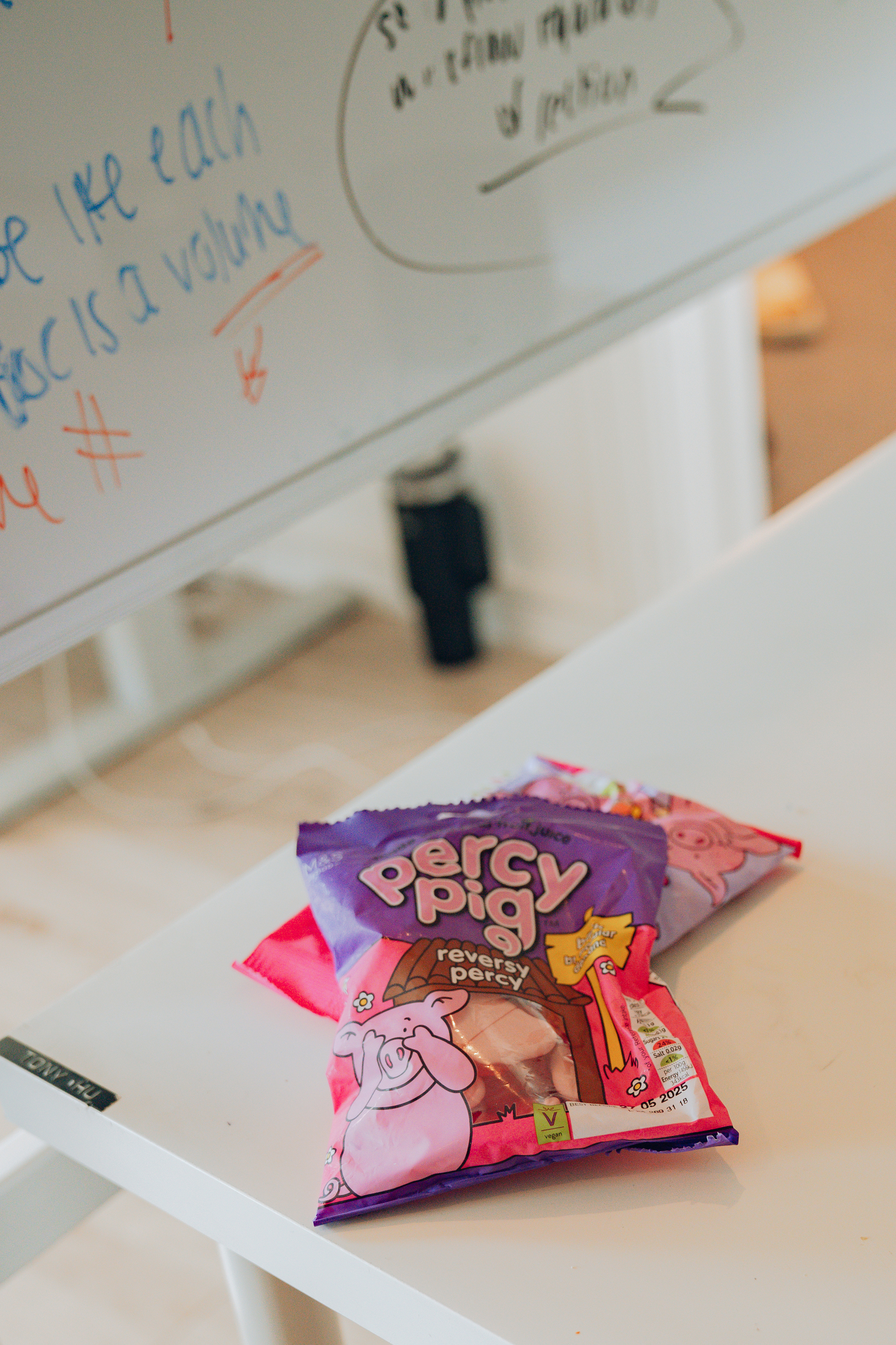 The image shows bags of Percy Pig candy on a white table near a whiteboard with colorful writing. The bags are brightly colored and prominently displayed.