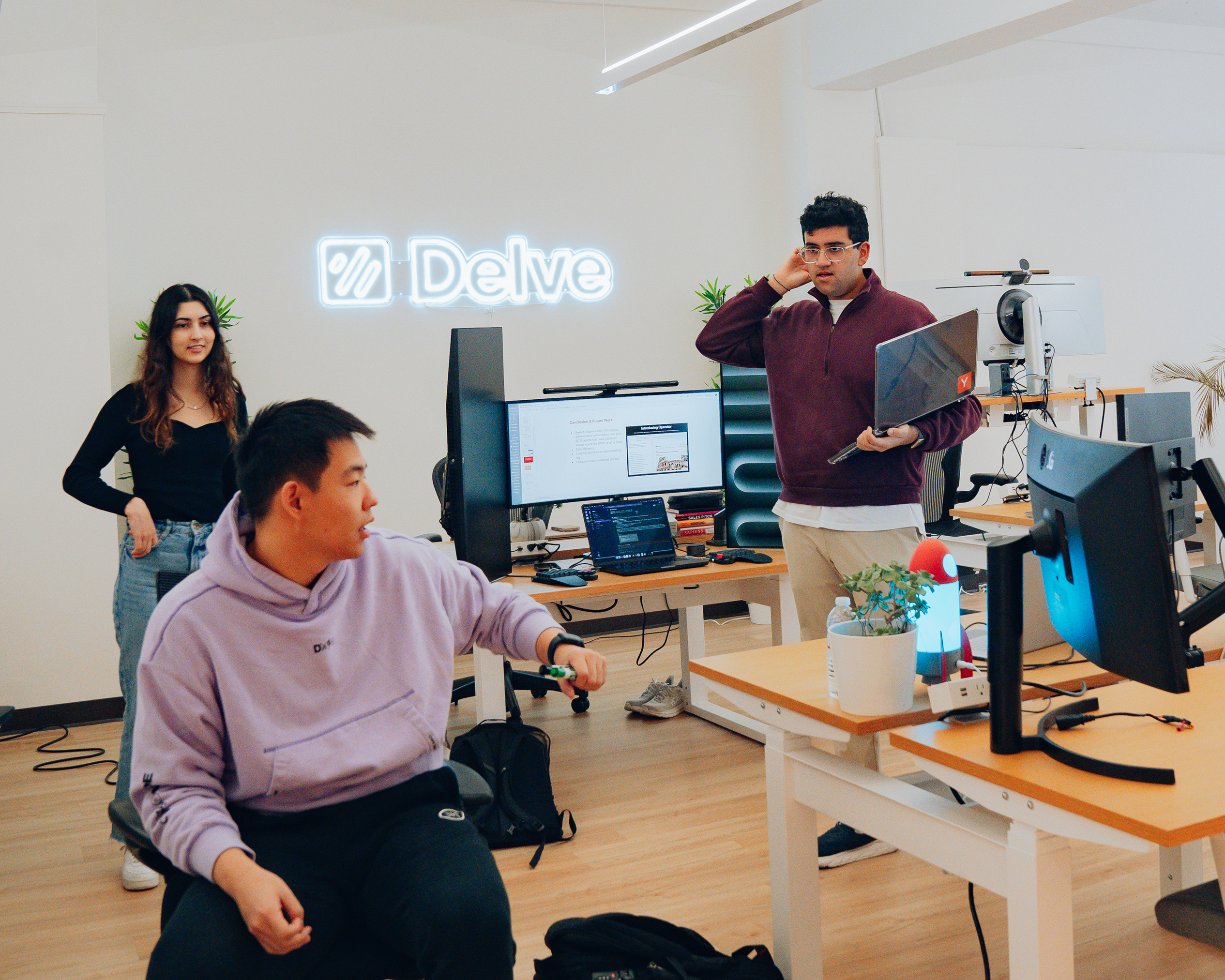 Three people are in an office with computers and a &quot;Delve&quot; sign. One is seated in a purple hoodie, another stands with a laptop, and a woman stands nearby.