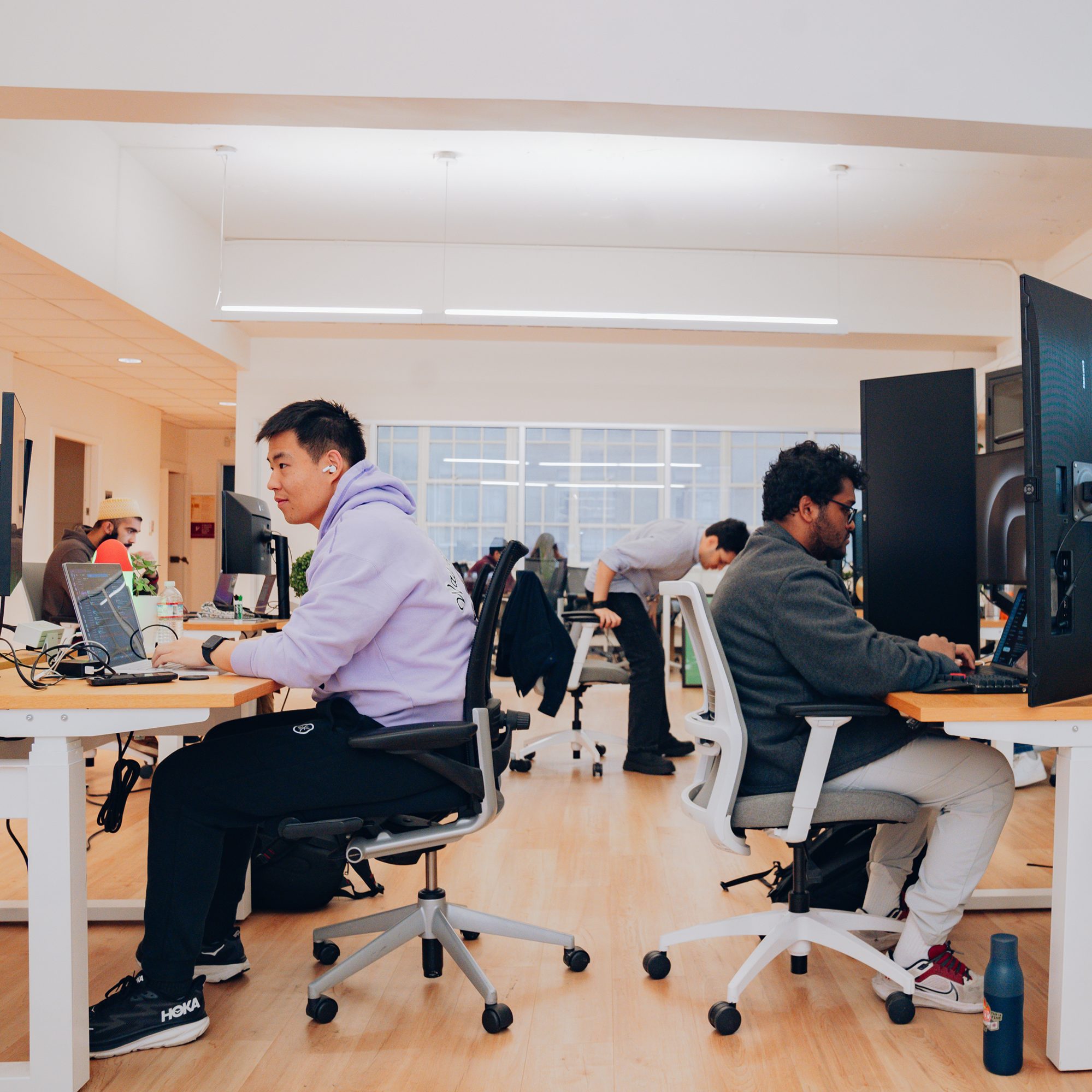 Two people are working at desks in an open office with computers. One wears a purple hoodie, the other a gray sweater. The room has bright lighting and light wood floors.