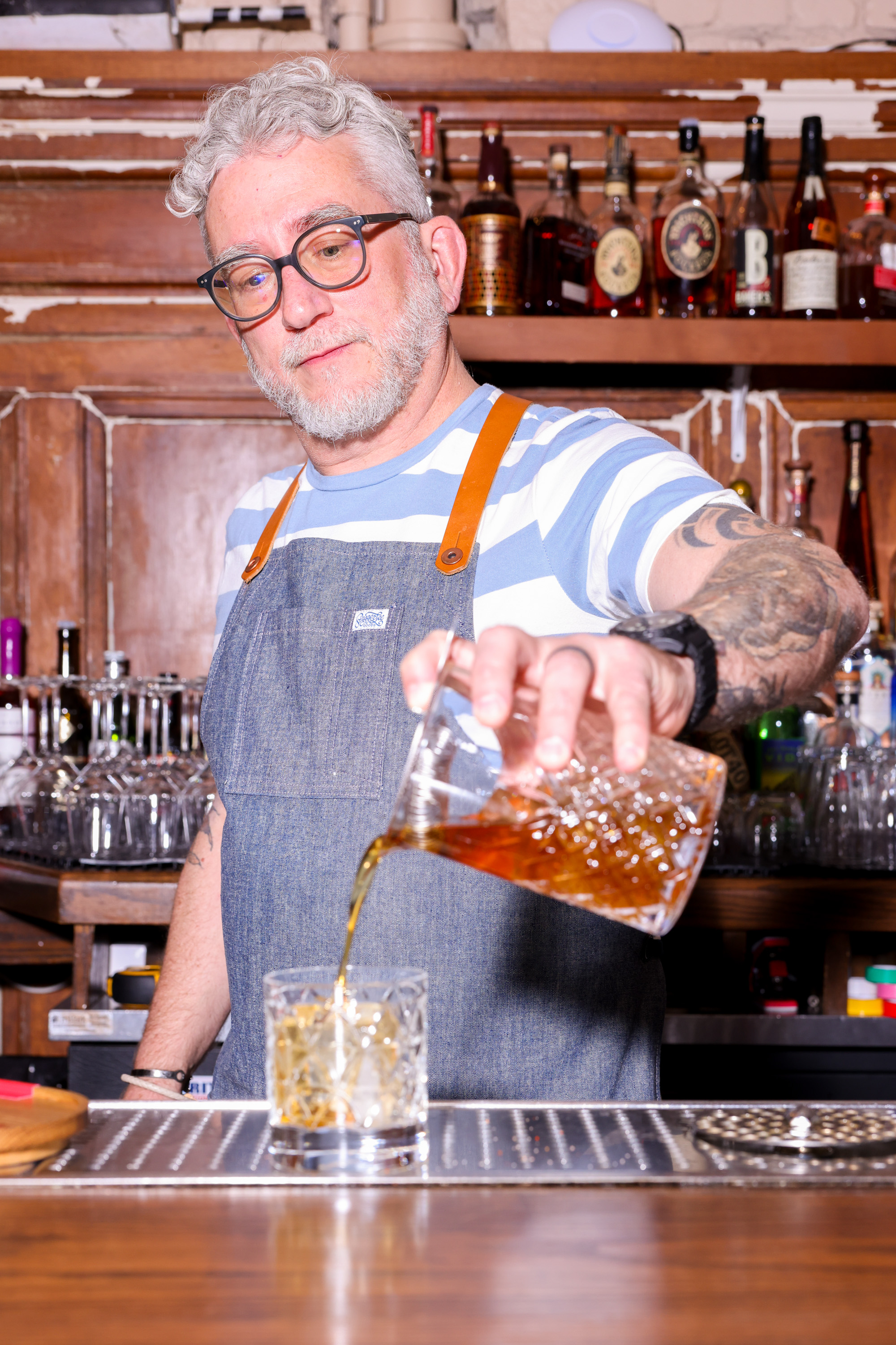 A bearded man wearing glasses and a striped shirt with a denim apron is pouring a brown liquid into a glass. Bottles are visible in the background.