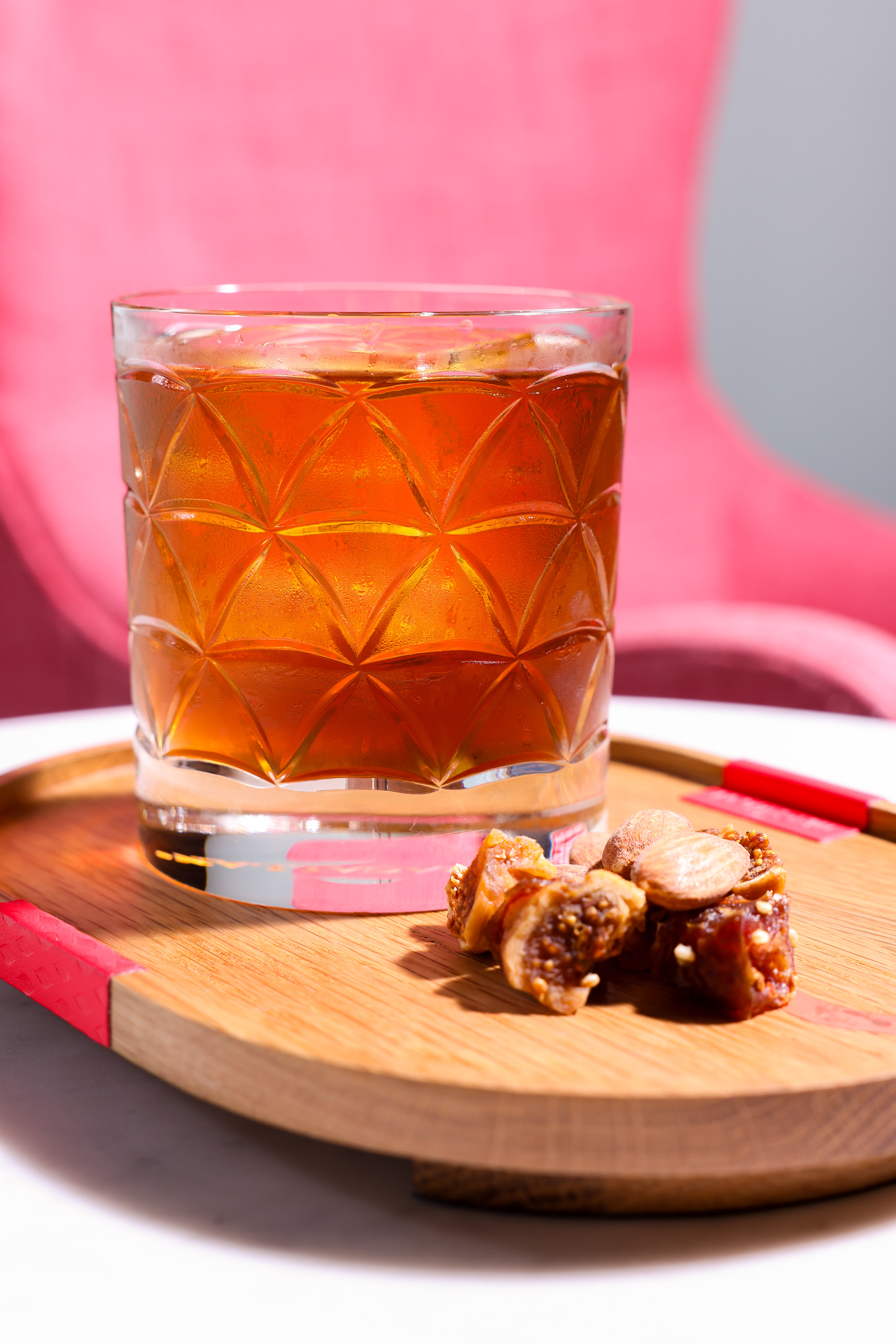 A patterned glass filled with amber liquid sits on a wooden tray next to dried fruit and nuts. The background features a bright pink chair.