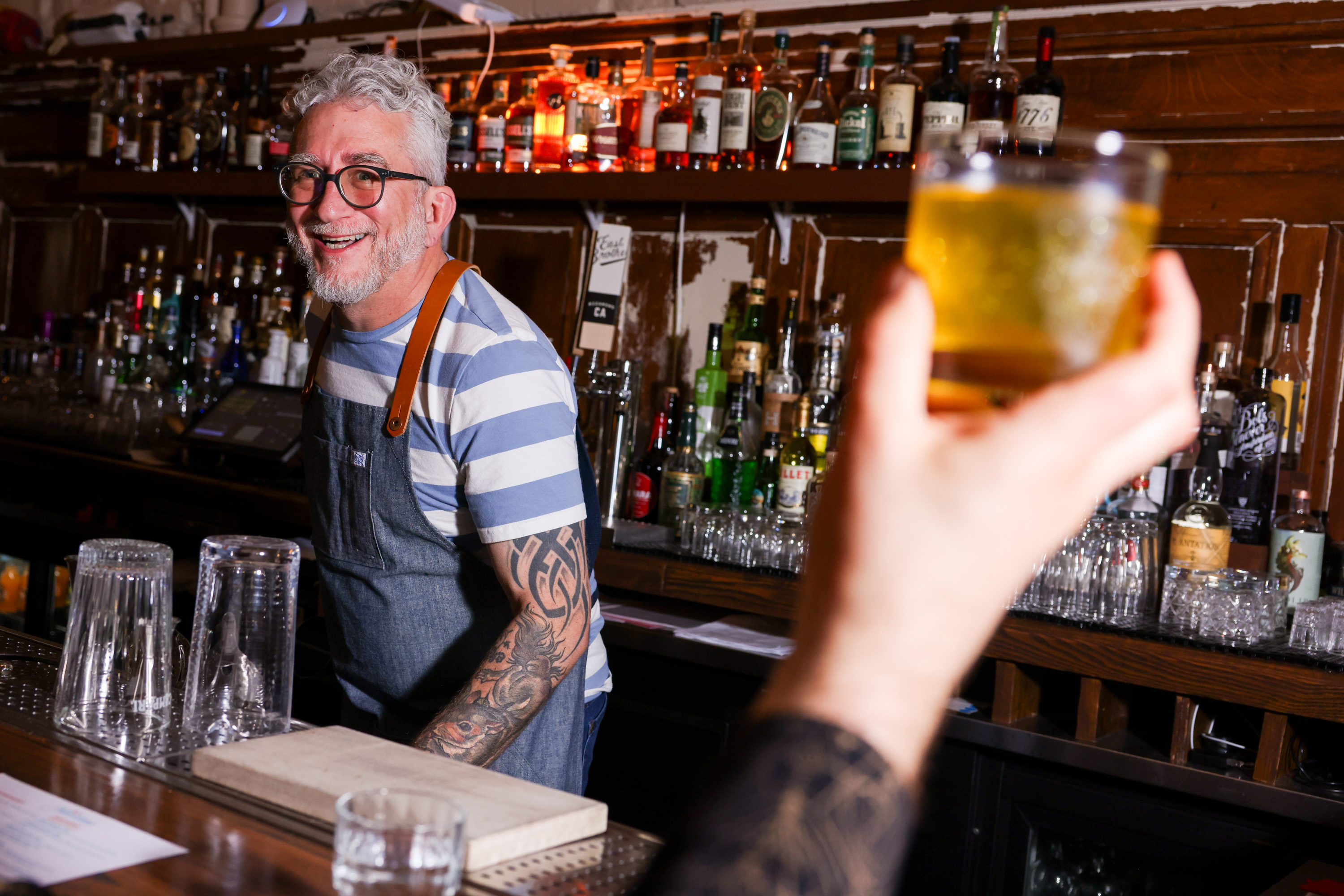 A smiling bartender with glasses and a tattooed arm stands behind a bar, wearing a striped shirt and apron. A hand in the foreground raises a yellow drink.