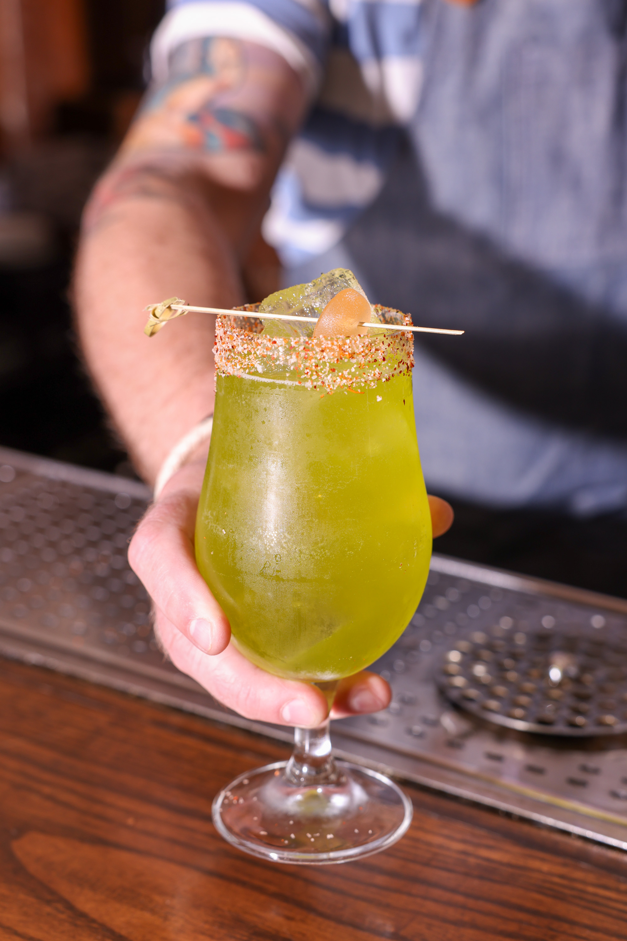 A person holds a green cocktail in a glass with a chili-salt rim, garnished with a fruit slice on a skewer. The background shows a metal bar surface.