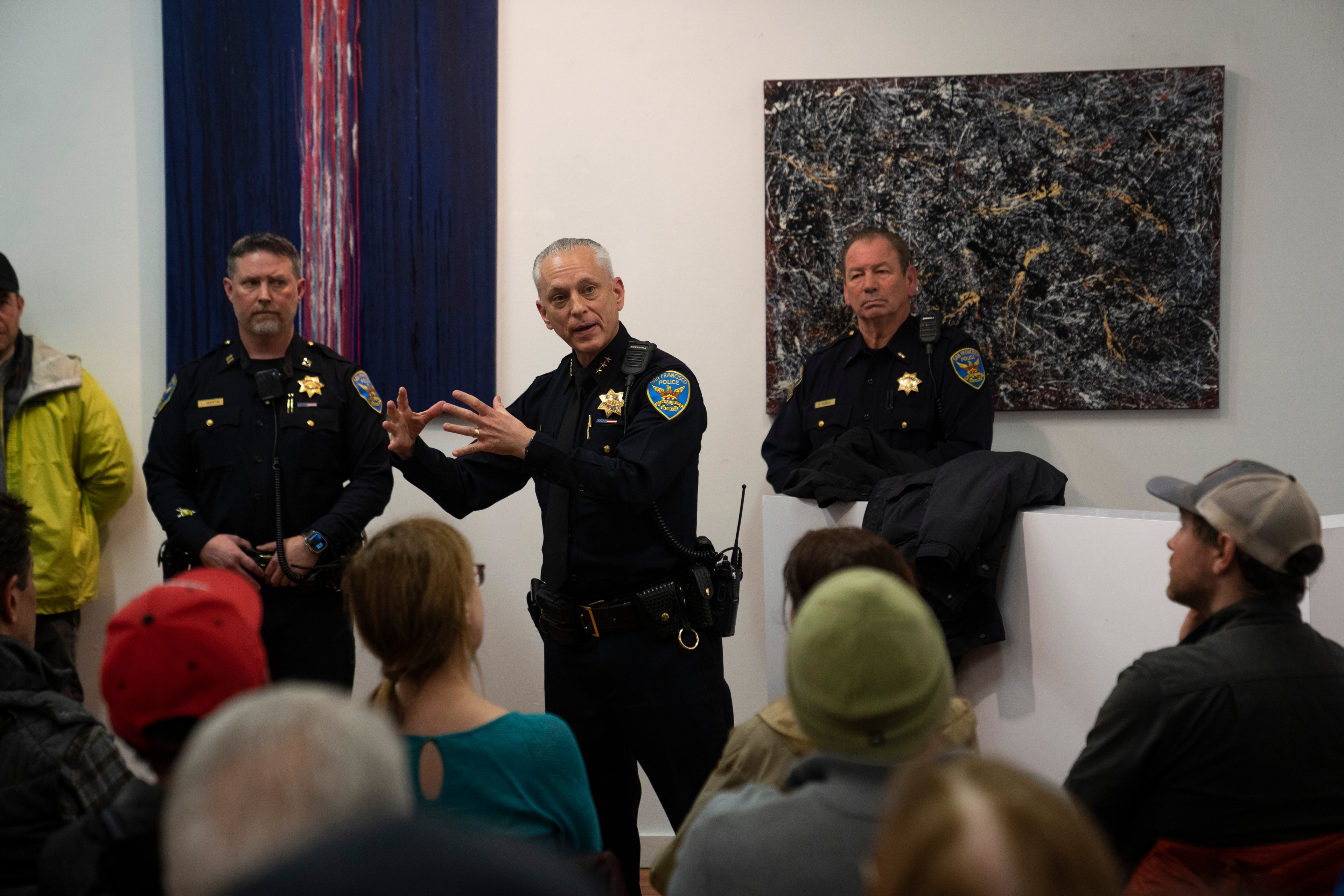 A police officer is speaking to a seated audience, gesturing with his hands. Two other officers stand nearby. Abstract paintings hang on the white walls behind them.