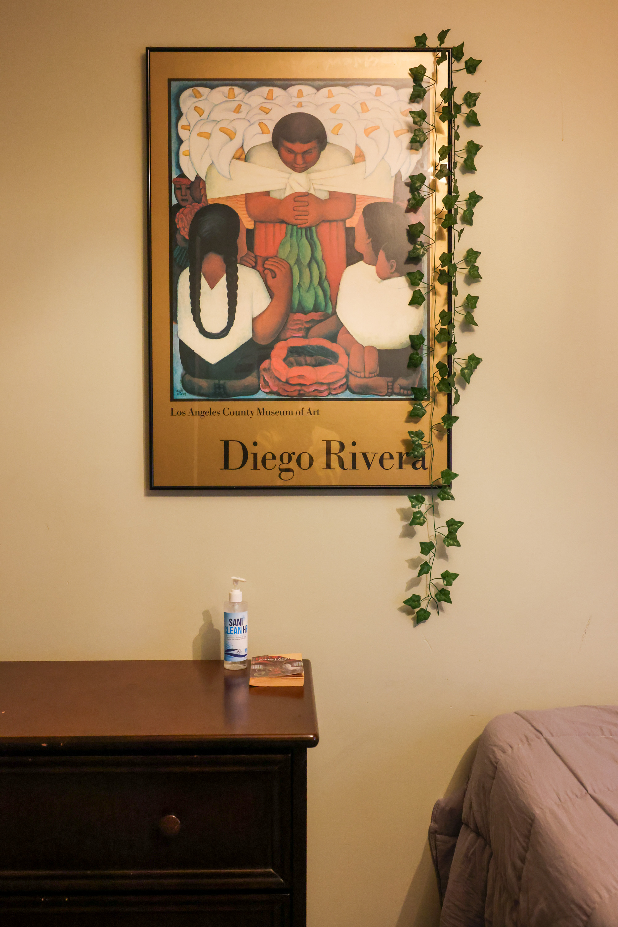 A gold-matted print of a Diego Rivera painting featuring three figres and white lily flowers hangs on the cream wall of a bedroom.