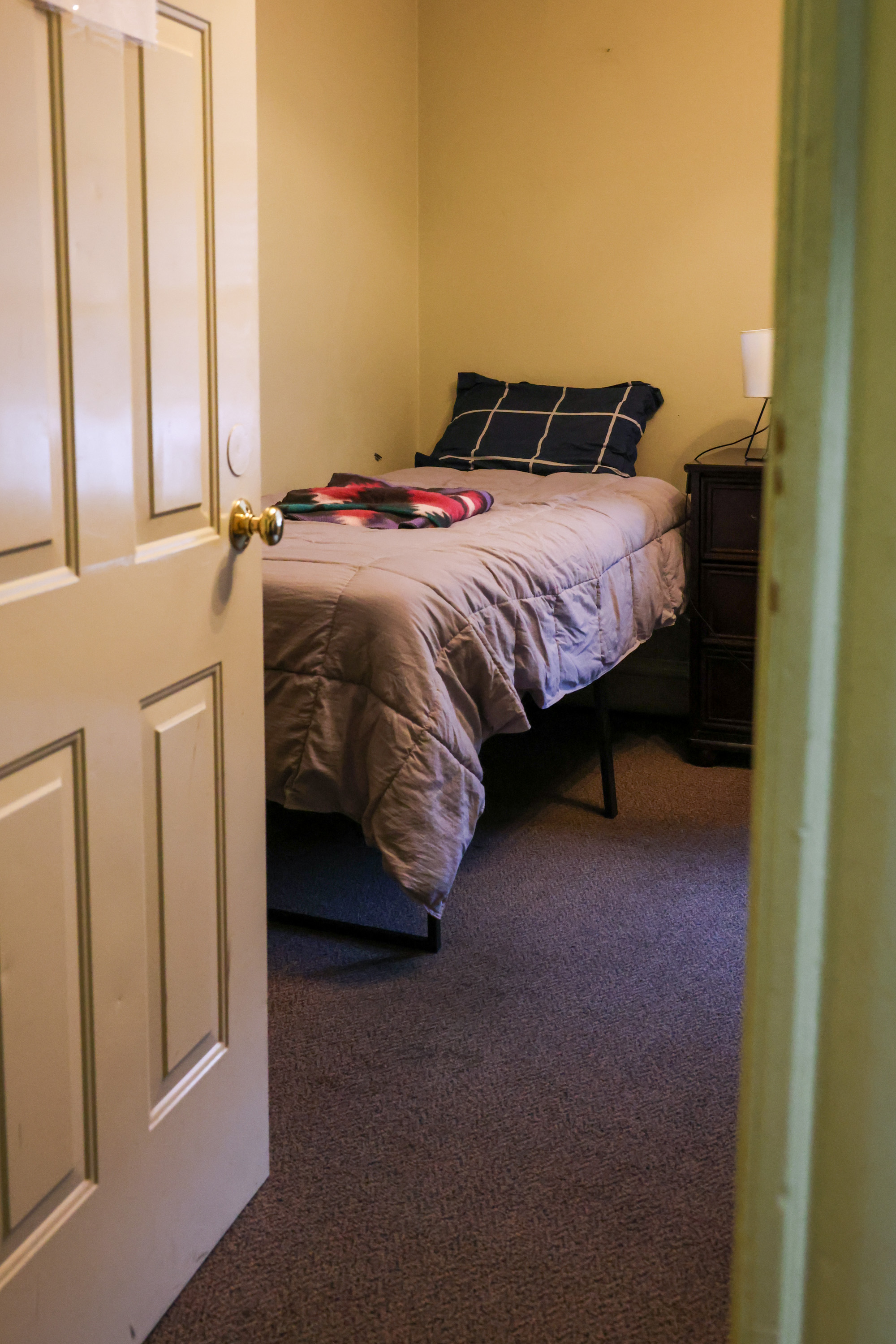 A twin bed with a gray comforter and dark plaid pillowcase is made neatly in the corner of a warm bedroom.