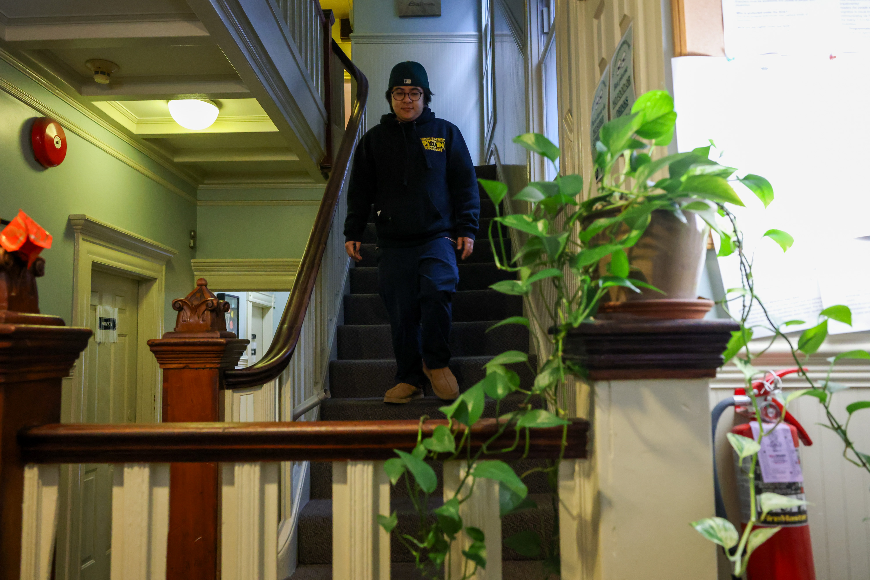 A person in a navy hoodie, hat and jeans walks down Edwardian style stairs decorated with a healthy philodendron.