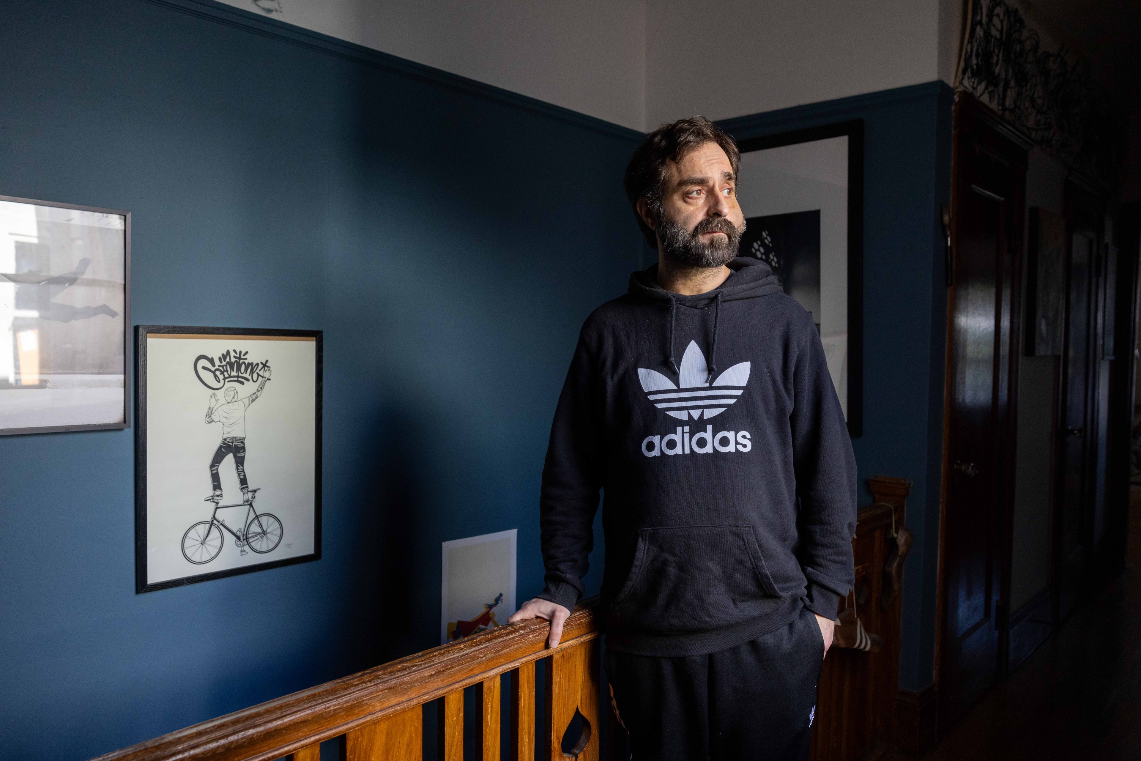 A man with a beard in an Adidas hoodie stands indoors by a wooden railing, beside framed artwork on a dark blue wall.