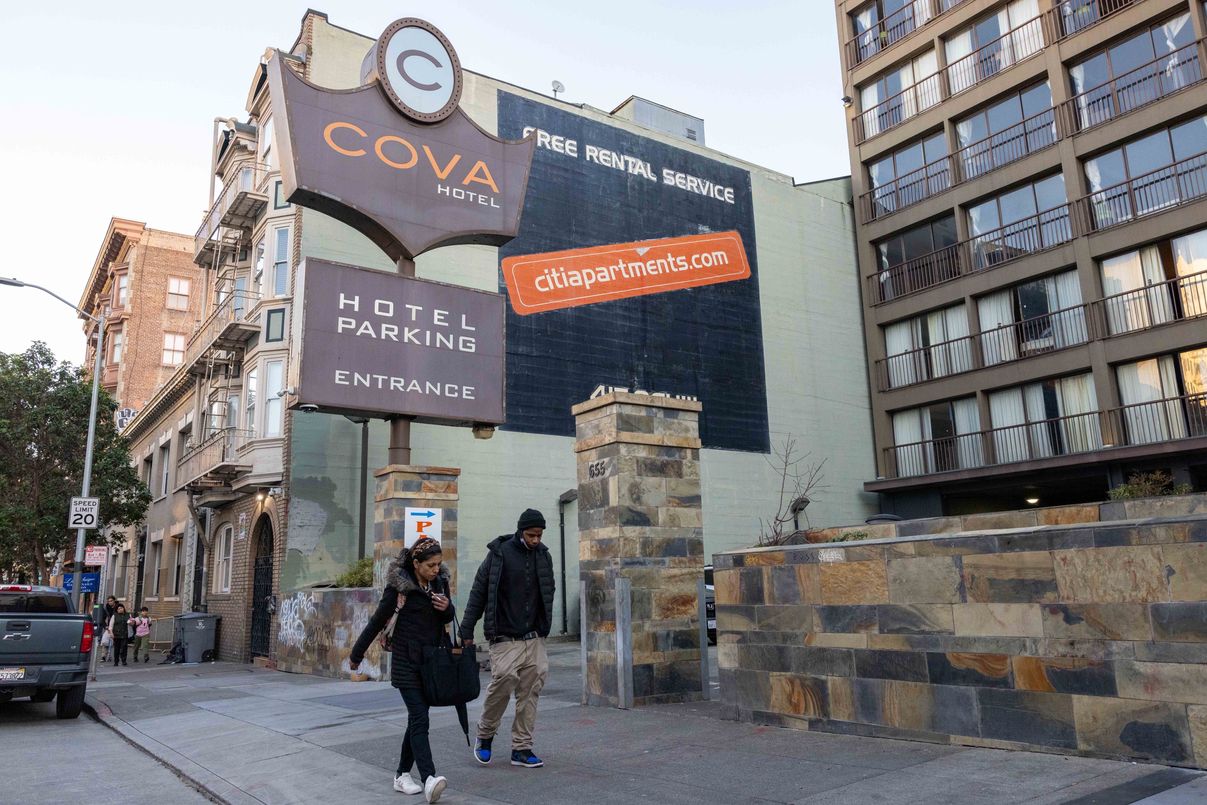 A couple walks past the Cova Hotel sign and a tall building. The scene is urban, with the hotel's advertisement, parked cars, and a speed limit sign visible.