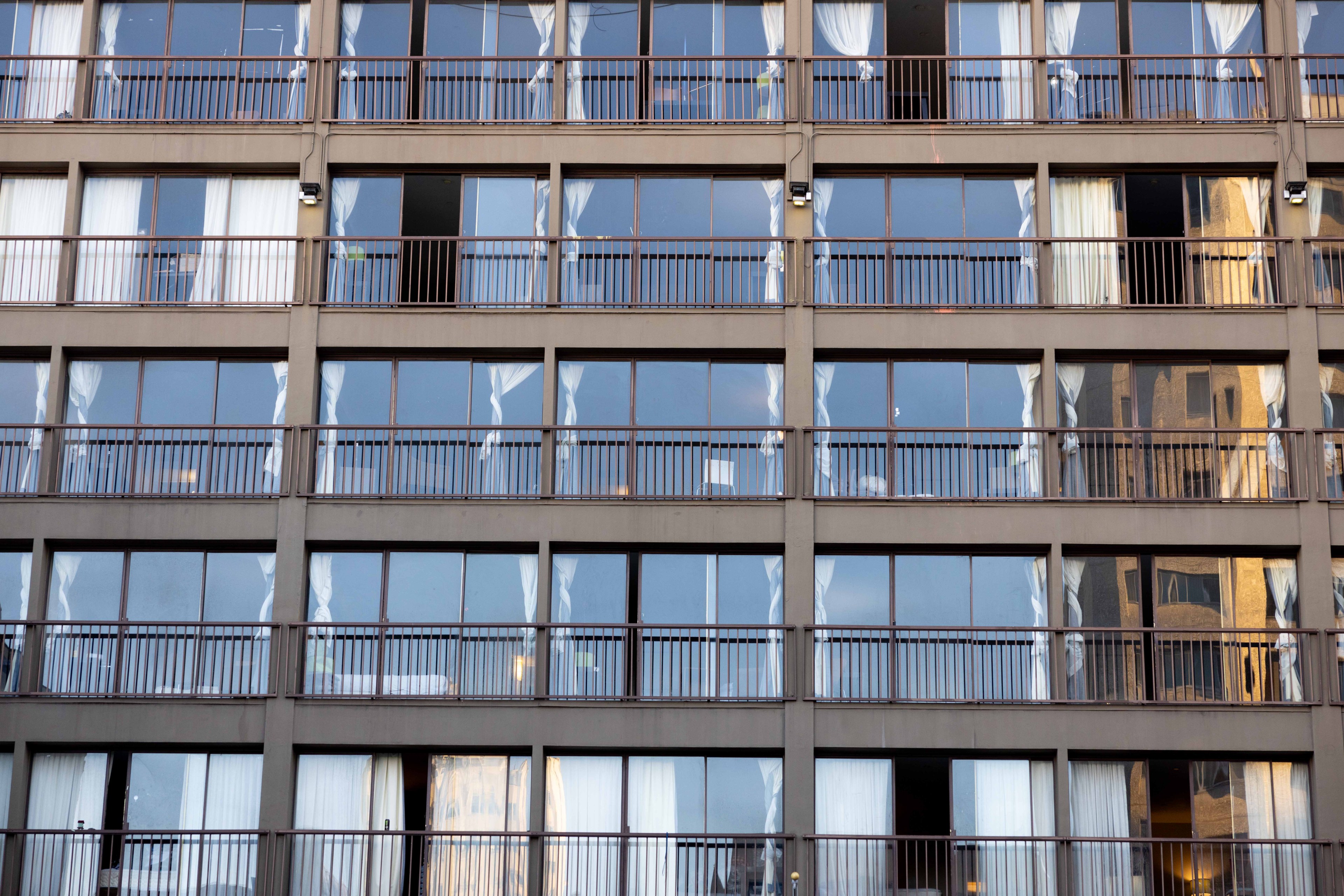 The image shows a multi-story building facade with rows of glass windows and balconies. White curtains can be seen through the windows, with some reflections visible.