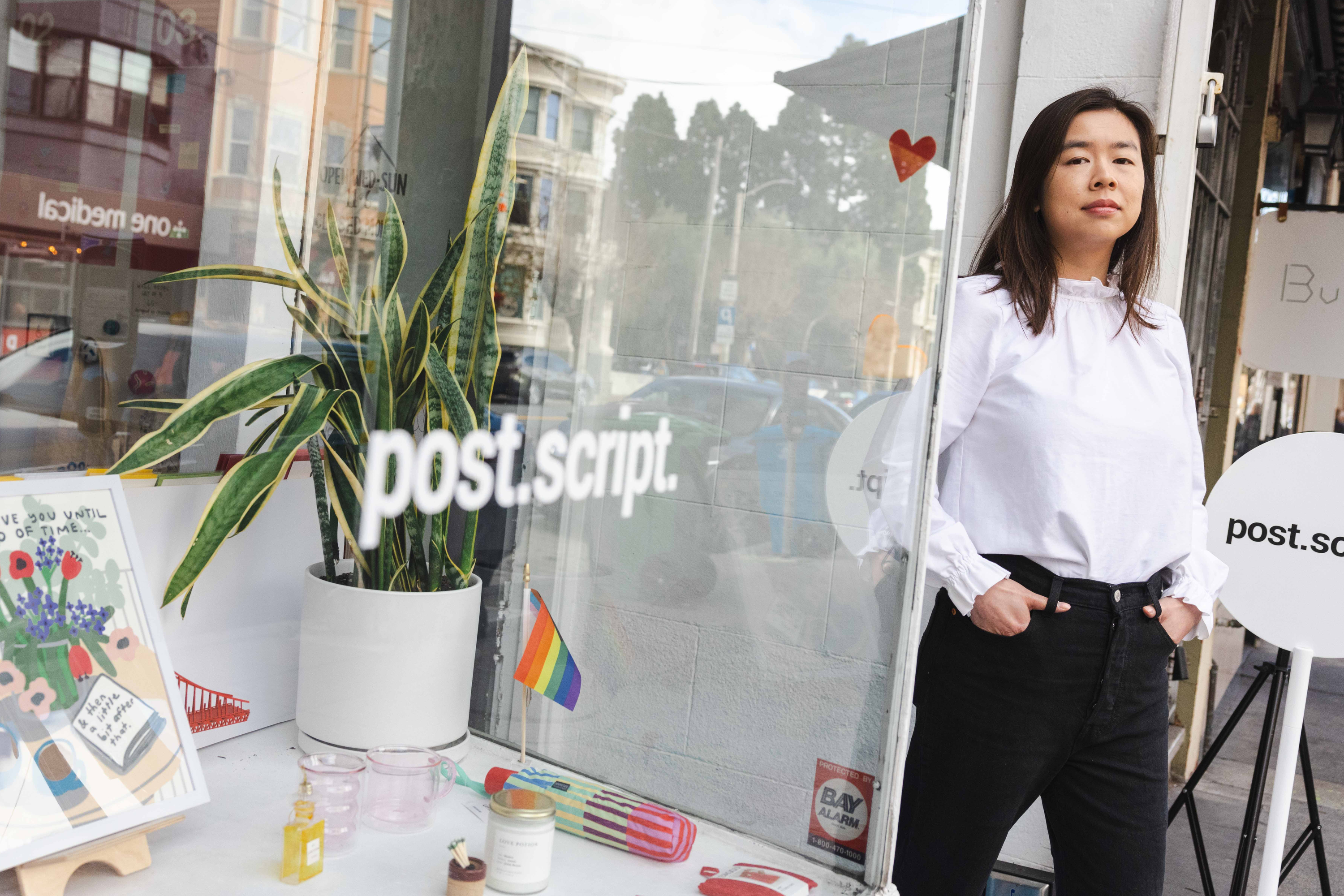 A woman in a white shirt stands outside a shop named "post.script." The window displays a plant, small items, a poster, and a rainbow flag.